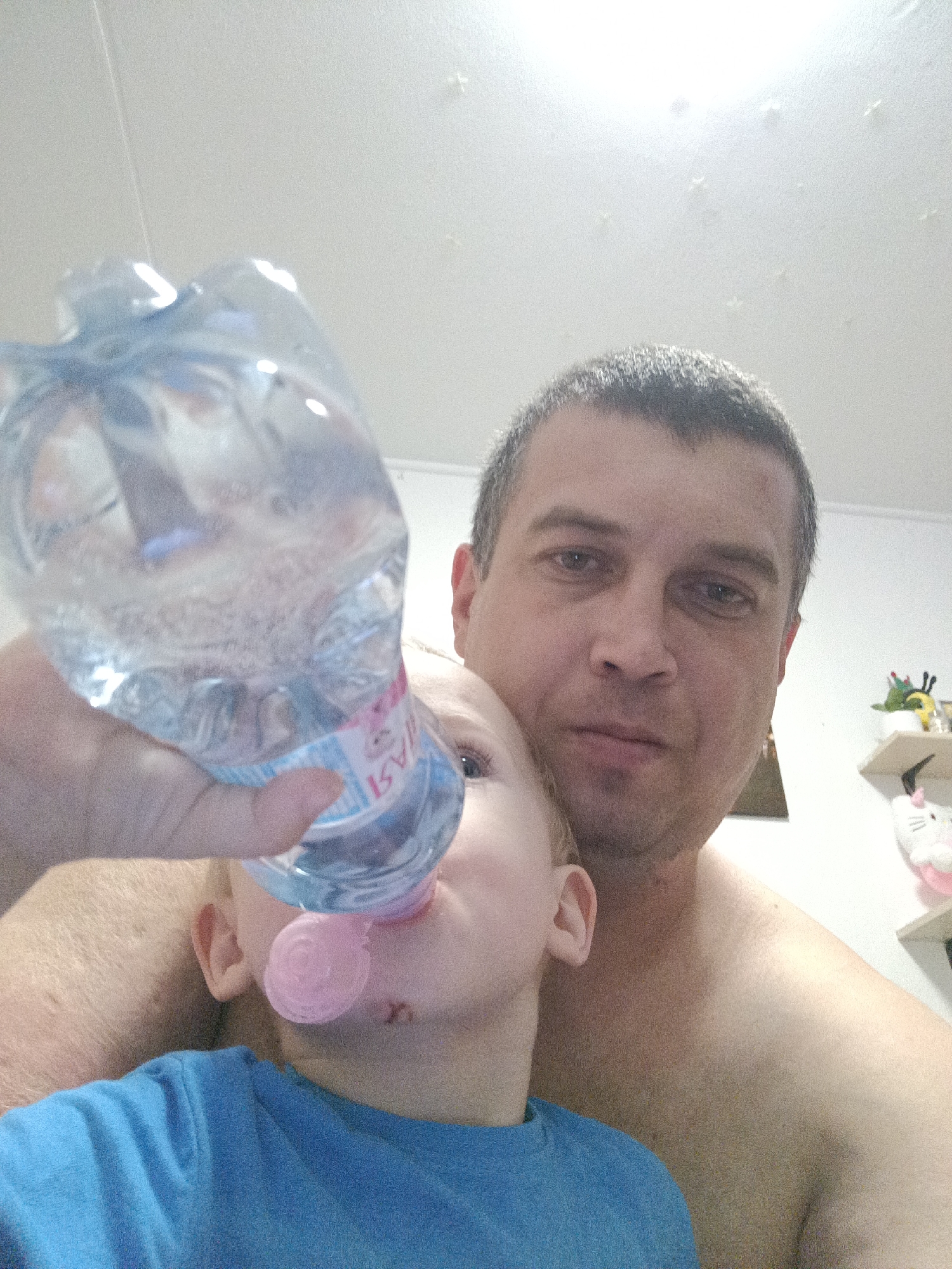 The image shows a man and a young child. The man is holding a bottle of water and is offering it to the child to drink. The child is wearing a blue shirt and has a pink pacifier in their mouth. The man is shirtless and appears to be topless. The scene is indoors, with a white ceiling and a shelf in the background that has some decorative items on it, including a stuffed animal. The lighting suggests it is daytime. The man has short, dark hair and is looking at the camera. The child is partially obscured by the man and the bottle. There is some text visible on the bottle label, which appears to be in Russian, with "AST" being partially legible. The overall mood of the image seems to be intimate and caring, depicting a moment of nurturing between the man and the child.