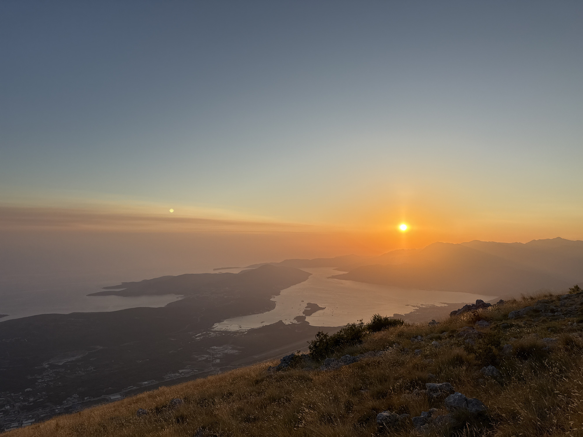 The image is a landscape photograph taken from a mountaintop overlooking a bay and a town. The sun is setting on the right side of the frame, casting a warm orange glow across the sky and water. The sky transitions from a clear blue at the top to a hazy orange near the horizon. A small, bright disc, possibly the moon or another celestial body, is visible in the upper left quadrant of the image.
The foreground consists of a grassy slope with dry, golden grass and scattered rocks. The midground shows the edge of a body of water, likely a bay or fjord, with undulating landmasses and faint outlines of mountains in the distance. The town, with buildings and roads, is visible along the coast and stretching inland. The overall atmosphere is serene and tranquil, with the fading light creating a sense of depth and mystery.
The location is a mountainous region in the vicinity of Kuk, Montenegro, likely offering a panoramic view of the Bay of Kotor or a similar coastal area. There are no people or animals visible in the image. The primary subjects are the natural landscape, the sunset, and the distant human settlement.