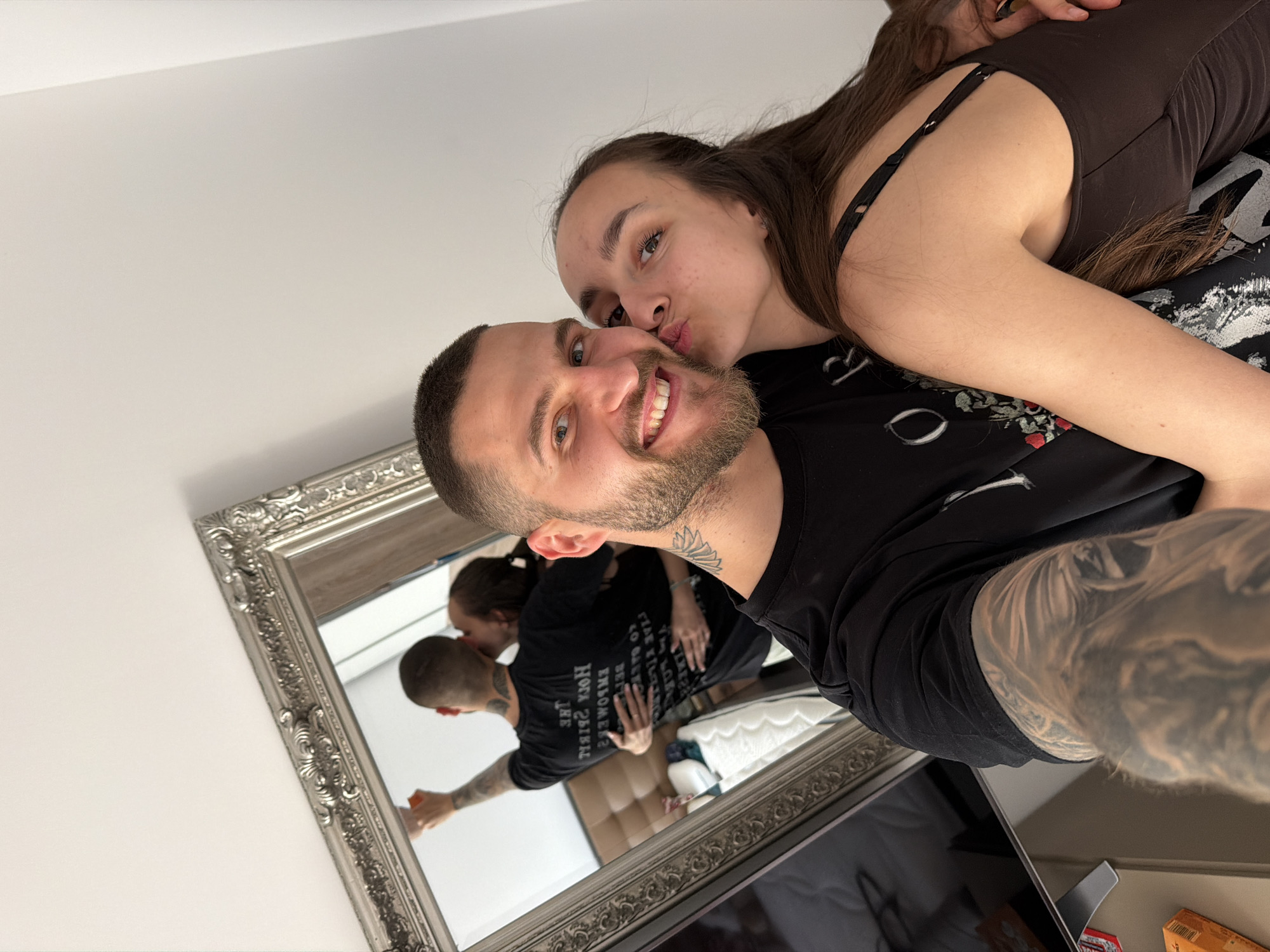 A tilted medium shot depicts a man and a woman in an embrace, with the woman kissing the man's cheek. The man, positioned in the foreground, sports a short haircut, a beard, and visible tattoos on his neck and arm. He is smiling at the camera. The woman, with long brown hair and wearing a brown tank top, is leaning over him. A large, ornate silver-framed mirror in the lower left corner reflects the scene, showing them in a similar embrace, but with the man appearing to clean the mirror. The environment appears to be an indoor space, possibly a bedroom, with a plain white wall visible in the upper left corner. Details discernible in the reflection include a bed with white sheets, a light-colored wall behind the couple, and hints of other objects on a table below the mirror. The setting is not definitively identifiable as Sofia, Bulgaria, lacking any characteristic features to confirm the location.