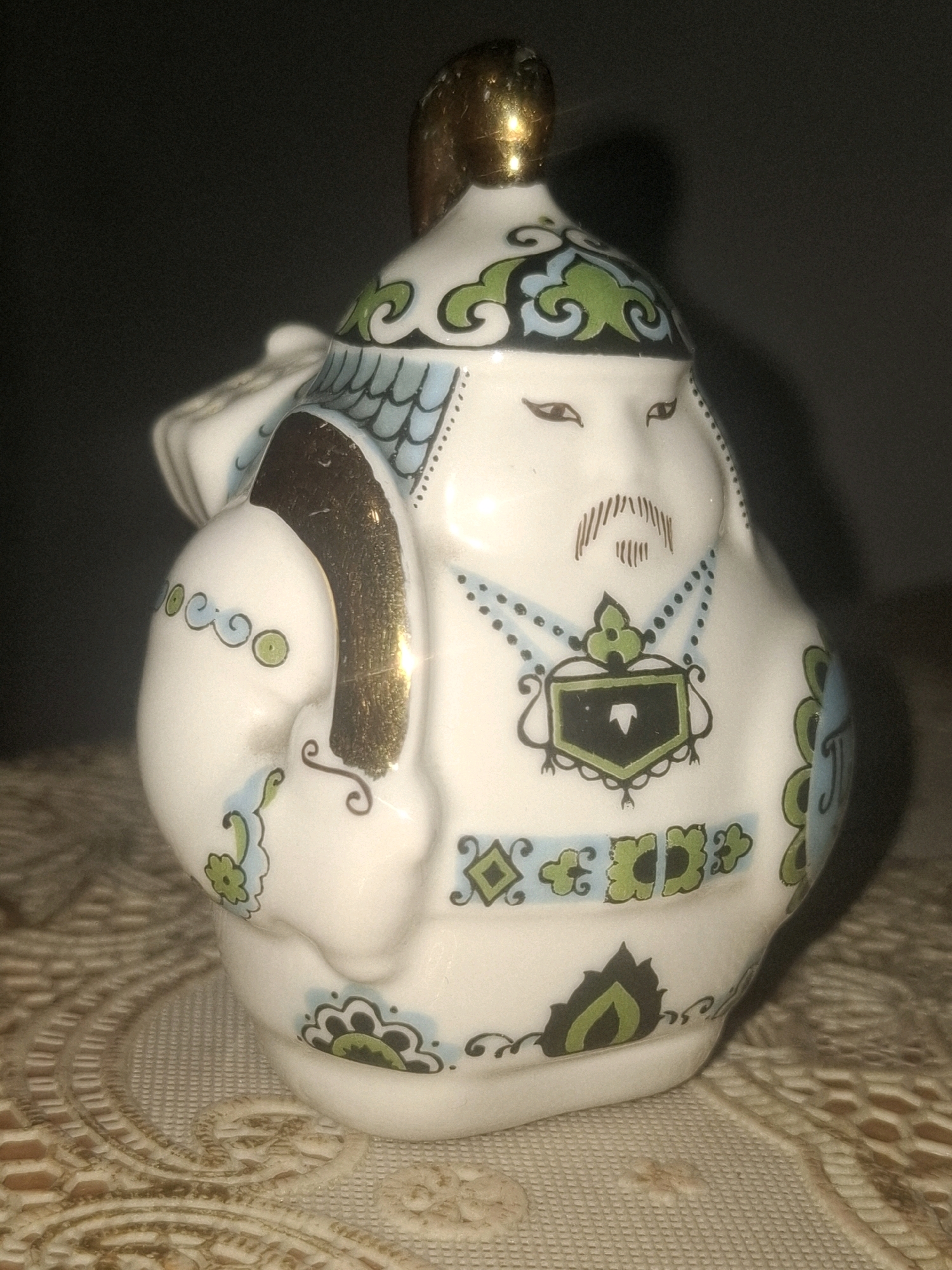 A porcelain figurine of a stout, bearded man in traditional attire sits on a decorative cloth. The figurine is white with blue, green, and gold accents. It has a gold-tipped headdress, painted eyes, and a thin mustache. The body is adorned with a patterned necklace and geometric designs. The figurine is depicted with one arm bent, holding something unseen. The background is dark and out of focus. The decorative cloth beneath the figurine has an intricate, embossed pattern in shades of beige and brown. There is no discernible connection to the city of Almaty, Kazakhstan.