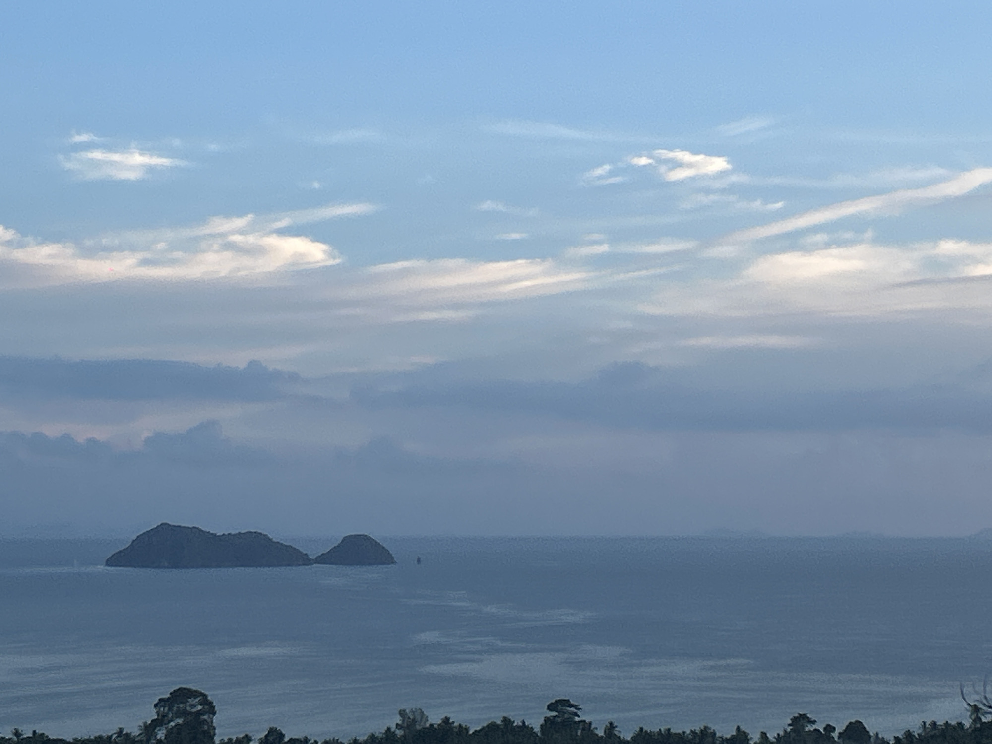 The image depicts a seascape with two visible islands in the distance. The sky is partially cloudy with varying shades of blue. The sea appears calm and dark. In the foreground, there are trees suggesting a coastal viewpoint or hillside. The location is ตำบลเกาะพะงัน, Thailand. No people are visible.