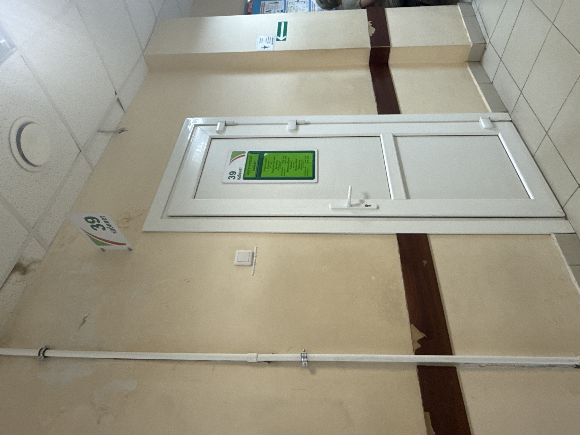 The image depicts a hallway in what appears to be a medical facility. The scene is indoors, likely in a building with a tiled ceiling and cream-colored walls. A prominent feature is a white door with a glass panel, likely a room entrance. Attached to the door is a green informational placard.
On the wall to the left of the door, a triangular sign with the number "39" is visible, suggesting a room number or office designation. Below this, a white pipe runs horizontally across the wall, attached by metal clips. Further down the wall, there are visible signs of wear and tear, including water stains and peeling paint.
Above the door, a white horizontal strip runs along the wall, possibly a trim or part of the building's structure. To the far left of the image, a circular vent or light fixture is visible on the ceiling. In the upper right corner, a section of a tiled wall is visible, with dark grout lines.
The lighting in the hallway suggests it is daytime, but there are no external windows visible to indicate the weather or specific time of day. The overall impression is one of a functional, though somewhat aged, interior space. There is no discernible activity or people in the frame. The environment appears to be clean but shows signs of age and maintenance needs in certain areas.
The placard on the door contains text in Cyrillic, which, along with the location context of Samara, Russia, suggests the text is in Russian. The sign with "39" also has Cyrillic text below it, likely indicating "кабинет" (cabinet/office).