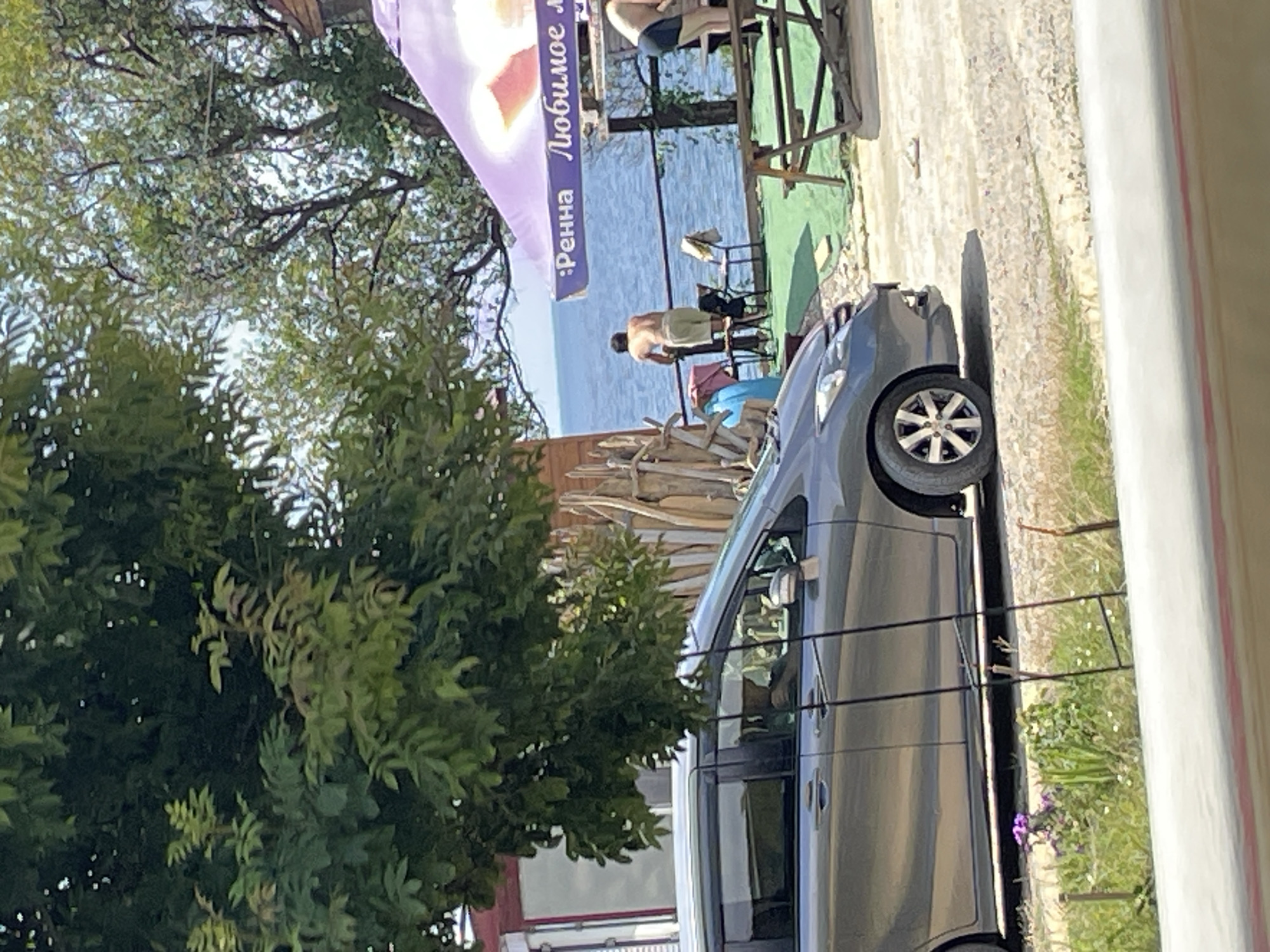 The image depicts a scene outdoors near the sea, likely in the city of Dzhubga, Russia. In the foreground, a gray minivan is parked on a gravel path. To the right of the minivan, a person is partially visible sitting in the driver's seat. Further back, near the sea, a man in light-colored shorts stands leaning on a railing, looking out at the water. Behind him, another person, wearing dark shorts, is also visible near the railing. A large purple umbrella with text that reads ":Ренна Любимое м" is open overhead. Several chairs and a wooden picnic table are situated on the grassy area near the water. Lush green trees and foliage are present throughout the scene, with one large tree dominating the left side of the frame. The sky is a bright blue, indicating sunny weather.