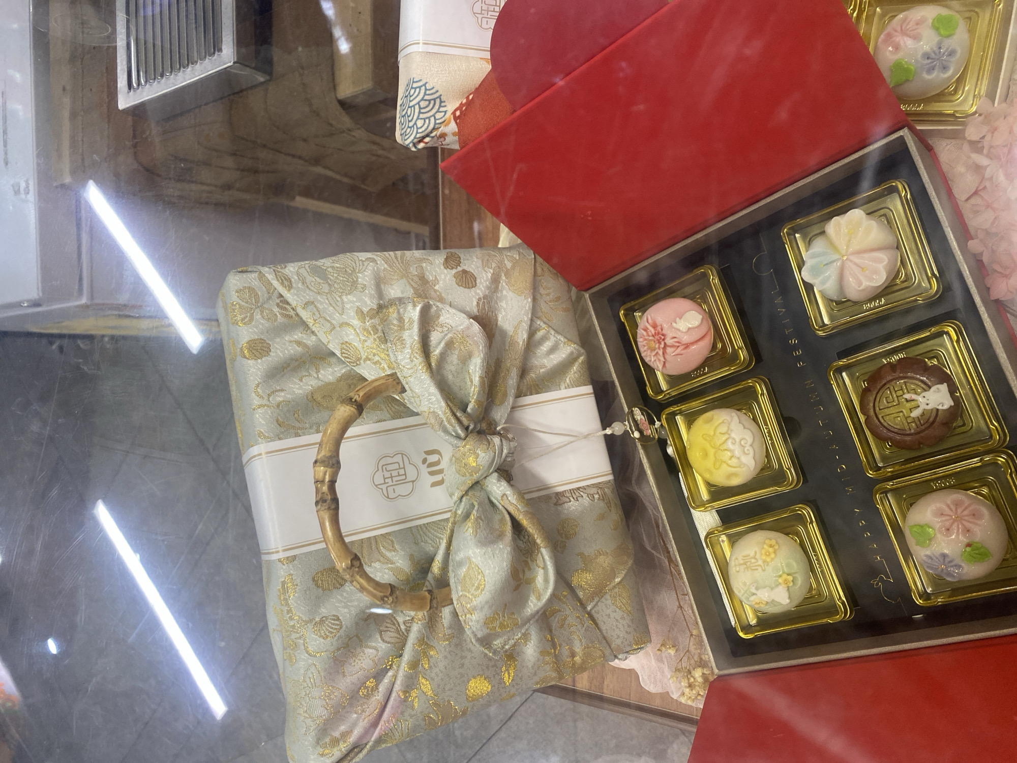 In Kuala Lumpur, Malaysia, a close-up, overhead shot captures a display of festive mooncakes within a retail environment, likely a bakery or gift shop. The scene is dominated by a large, elegantly wrapped gift box in the upper portion of the frame. This box is adorned with a light green fabric featuring an intricate gold floral pattern, secured with a large bow and a natural bamboo handle. A white band with East Asian calligraphy and a gold emblem is wrapped around the gift.
Below this, a red gift box opens to reveal several individual mooncakes arranged in gold square molds. These mooncakes display a variety of decorative designs. One is a pale pink with a floral motif and a small rabbit figurine. Another, to its right, is pale yellow with an elaborate pattern of rabbits and clouds. A third mooncake is a light green, also decorated with flowers and a rabbit. To its right, a fourth mooncake is a pastel assortment of pink, blue, yellow, and white, arranged like petals. In the lower row, a dark brown mooncake is embossed with a circular pattern and a rabbit. Next to it is a round, light pink mooncake with floral decorations and a rabbit. The words "HAPPY MID-AUTUMN FESTIVAL" are faintly visible printed on the dark surface of the open red box.
The background shows a reflective surface, possibly a glass display case, with overhead fluorescent lighting creating bright streaks and reflections. A metallic grate or vent is visible in the upper right, and some wooden shelving or display elements are present. The overall impression is of a well-presented collection of traditional Mid-Autumn Festival treats.