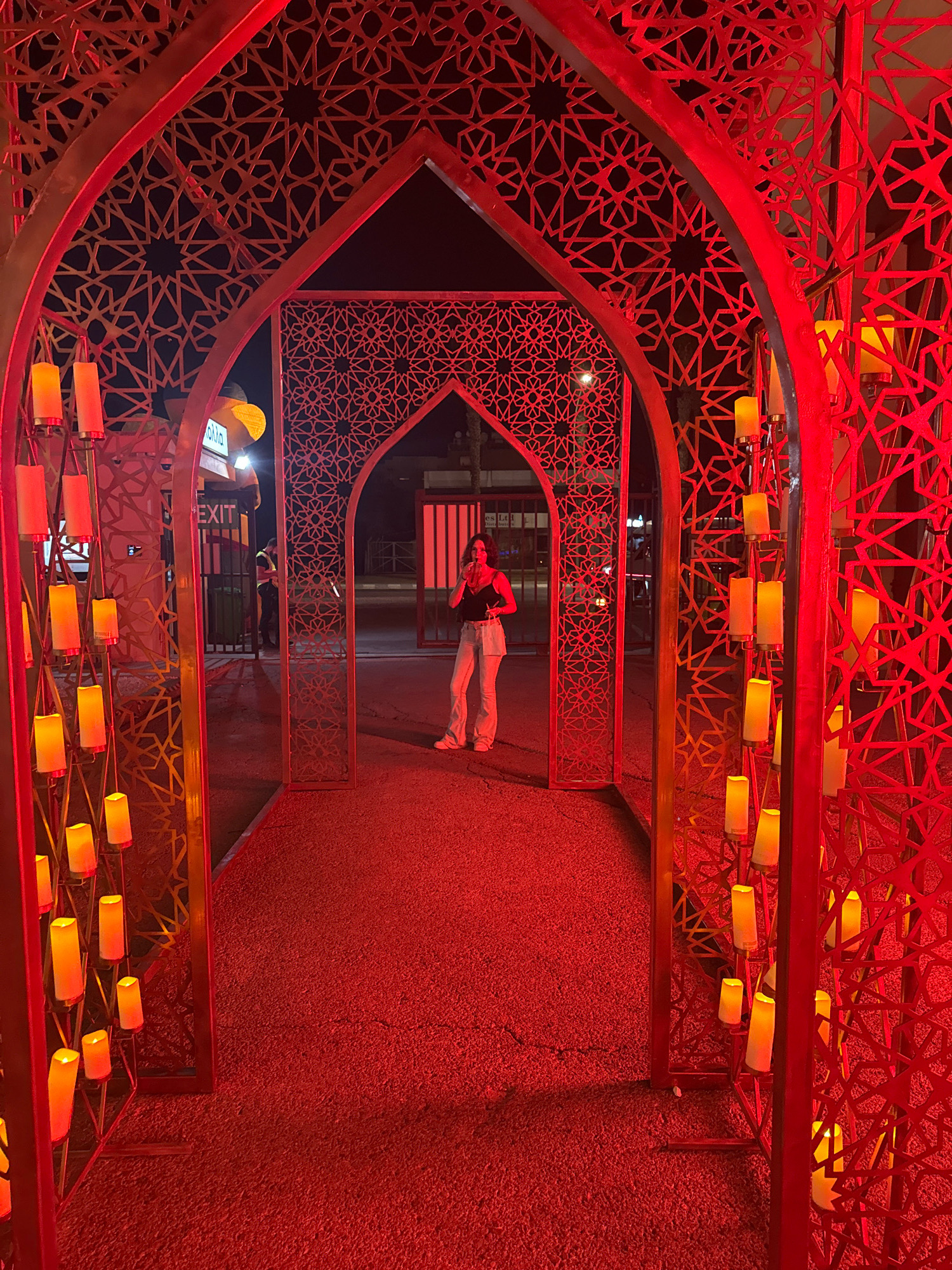 An outdoor scene at night, illuminated by red lighting and decorative orange-yellow lights resembling candles. The foreground features a pathway made of red gravel leading through a red metal archway decorated with intricate geometric patterns. To the left, a vertical structure displays numerous cylindrical lights.
In the middle ground, a young woman with dark hair is standing, holding a drink to her mouth and looking towards her left. She is wearing a black tank top and light-colored wide-leg pants. Behind her, an "EXIT" sign is visible on a building. There are also people in the background, including a figure in an orange vest and another in a dark uniform. The setting appears to be a public space, possibly an event or venue, with a street and buildings visible in the distance. The overall ambiance is festive and theatrical. The location is identified as Limassol, Cyprus.