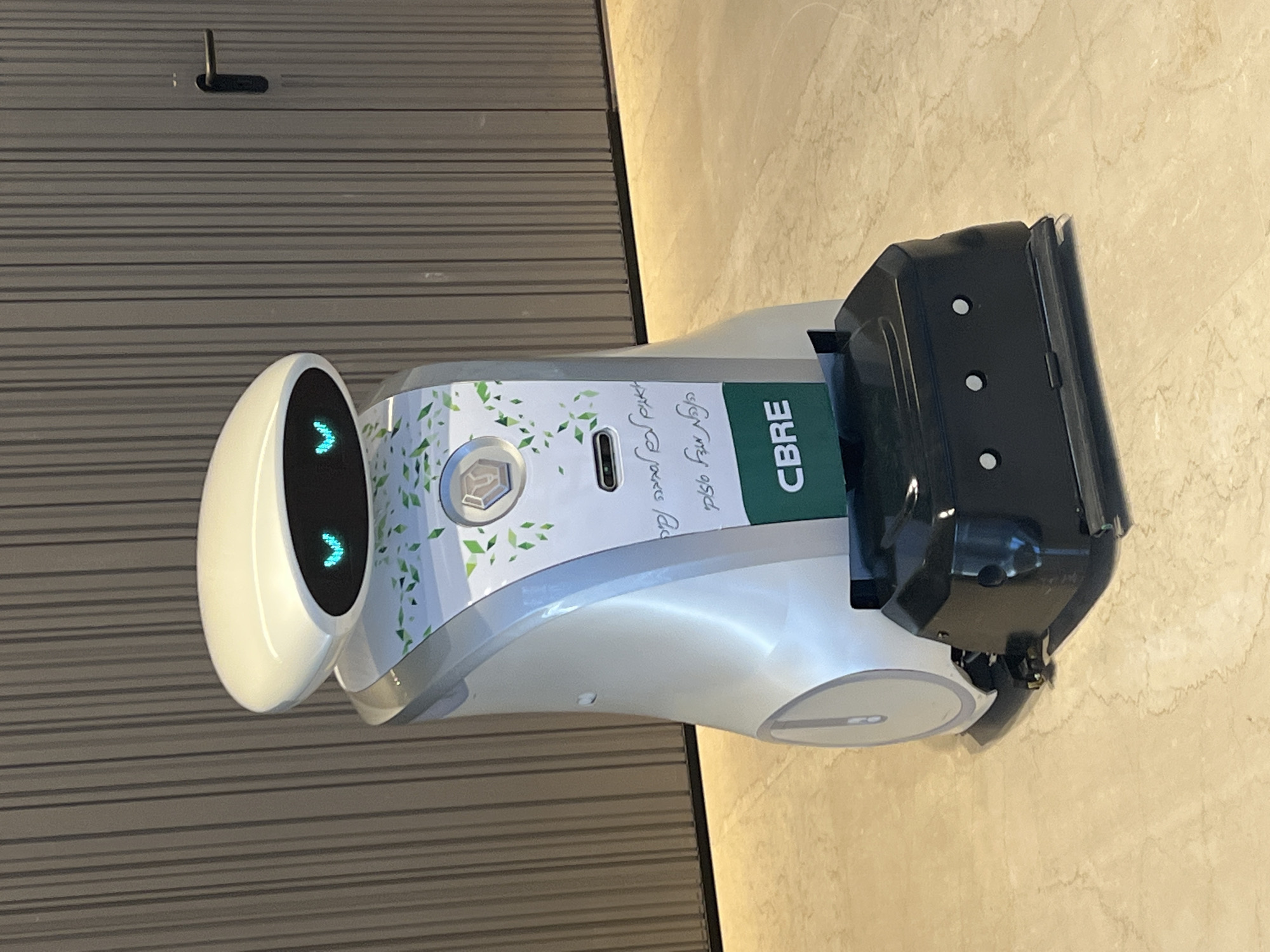 The image displays a modern service robot standing indoors, likely within a commercial or office building in Tel-Aviv, Israel. The robot features a sleek, rounded design with a white and silver chassis. Its 'head' is a black, oval screen displaying two glowing blue chevron shapes, which resemble eyes or directional indicators.
The robot's central body section is decorated with a white and light green graphic wrap, featuring a pattern of small leaves. Vertical green text on this section clearly reads "CBRE", identifying a brand or company. Below the leaf pattern and above the CBRE text, there is handwritten text in Hebrew script, which appears to be a holiday greeting such as "חג כשר ושמח" (Happy and Kosher Holiday).
Attached to the robot's rear is a black, robust-looking container or dispenser with visible circular perforations. The robot is situated between two distinct walls: a dark grey wall with horizontal striped paneling to its left, and a lighter, mottled, possibly marble or stone-textured wall to its right. A small, black, L-shaped fixture or hook is mounted on the striped wall above the robot's head. The robot appears to be stationary, perhaps in a waiting or operational stance.