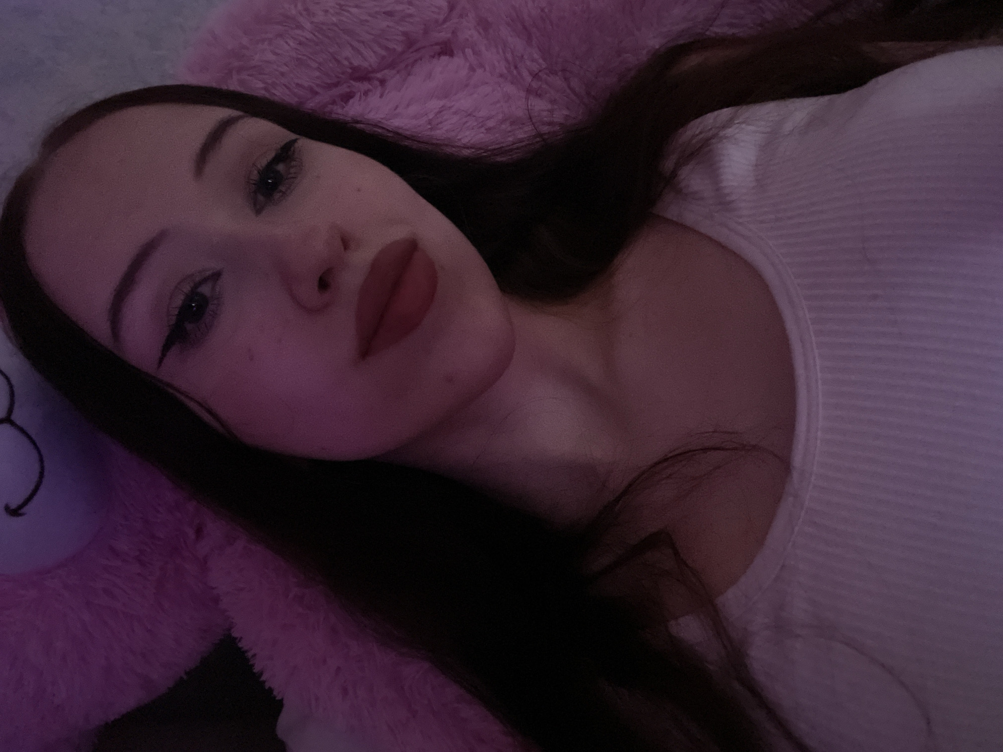 The image is a close-up, selfie-style portrait of a young woman lying down. She has long, dark hair and is wearing a white ribbed tank top. Her makeup includes winged eyeliner and matte red lipstick. She is positioned against a soft, fluffy pink surface, which appears to be a large stuffed animal or blanket. The lighting is dim and tinted pink, creating a soft glow. There is no discernible information about the location within Votkinsk, Russia, or any specific environmental details that would place it within the city. The scene is a personal, indoor setting.