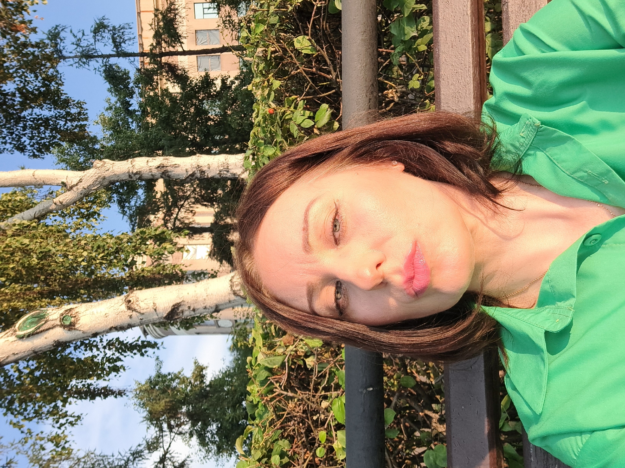 A woman is sitting on a park bench outdoors, in front of a dense green hedge and several trees. She is wearing a green collared shirt and a gold necklace. Her hair is brown and shoulder-length. She is looking directly at the camera with her lips pursed, as if blowing a kiss. The sunlight is hitting her face from the right side of the frame, creating a warm glow. In the background, there is a building and the sky. The image appears to be taken during the daytime, likely in the late afternoon or early evening, given the quality of the light. The location is the city of Irkutsk, Russia.