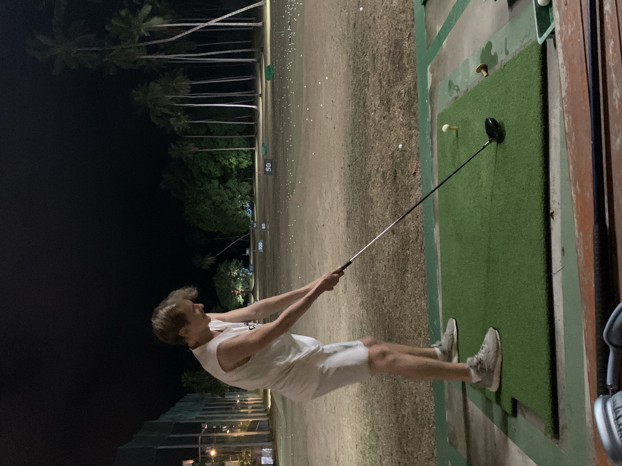 A male golfer is swinging a golf club at a driving range in บ้านบางรักษ์, Thailand. He is wearing a white tank top and shorts, and grey sneakers. The driving range has palm trees in the background, and yardage markers. A golf ball is on a tee on a green mat. Used golf balls are scattered on the ground in the distance. It is night.