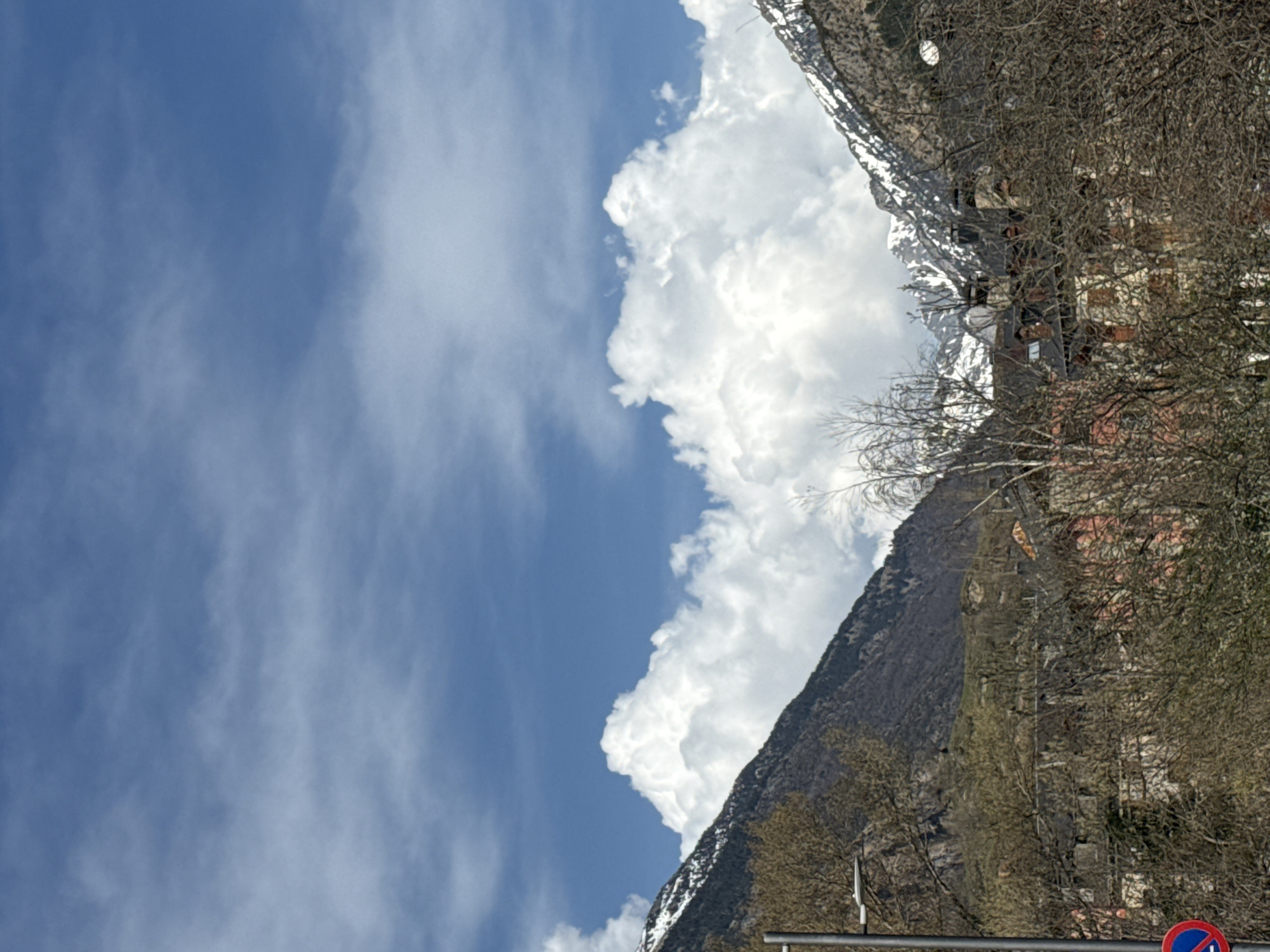 The image shows a view in the city of Benasque, Spain. A clear blue sky with wispy clouds fills the left portion of the frame. Fluffy white clouds contrast against the sky near a mountain range that rises in the background. Patches of snow are visible on the higher elevations of the mountain, which transitions into rocky terrain further down. The lower slopes of the mountain are covered in trees. Buildings are partially visible among the trees and vegetation on the right side of the frame. Bare branches are visible in the foreground. A no parking sign is also partially visible.