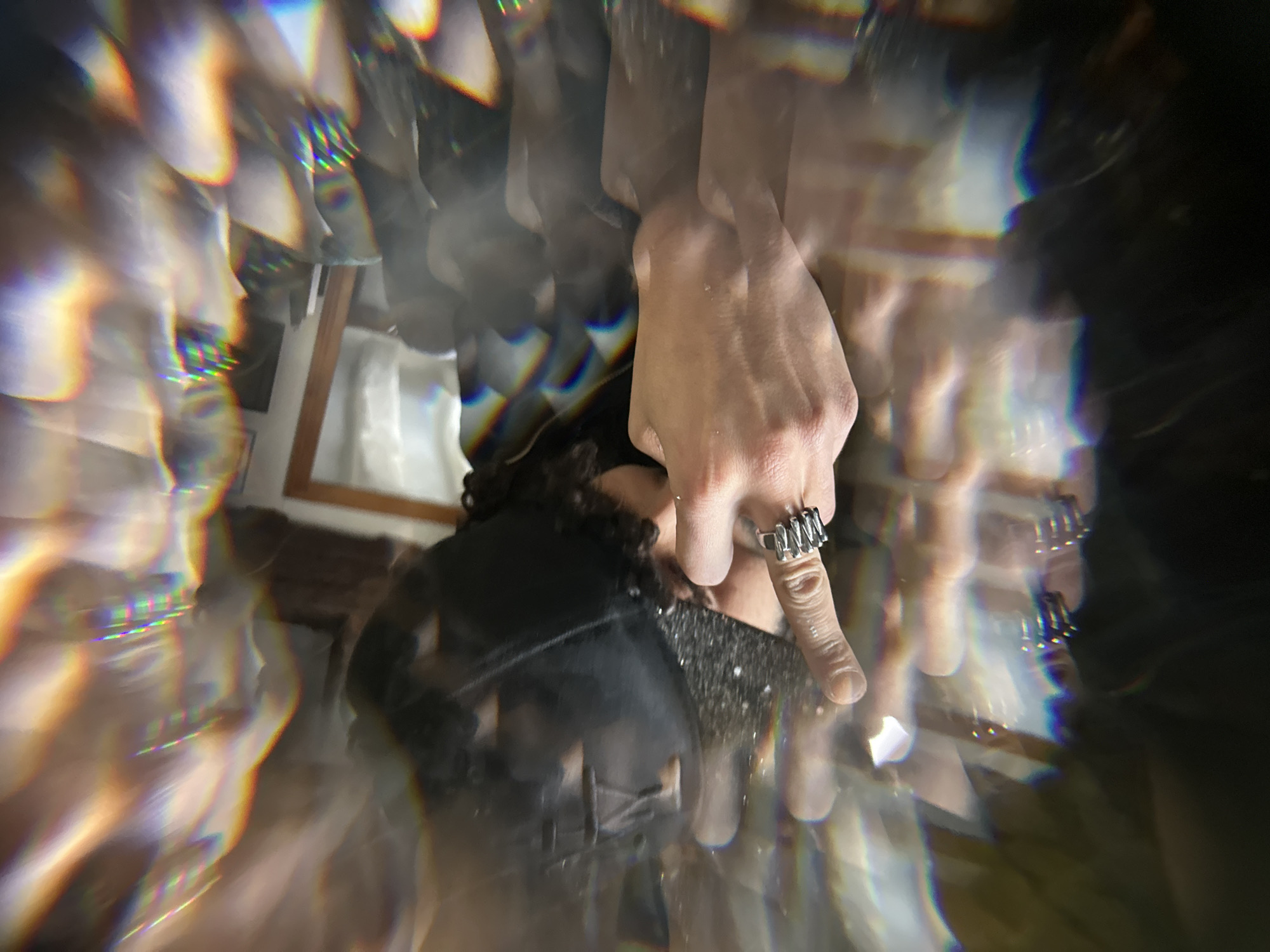 The image presents a distorted view, likely through a prism or lens, of a person wearing a black top with what appears to be a sparkly embellishment on the collar. Their left hand is prominently visible, adorned with a silver ring on the index finger. The background is partially discernible, revealing what seems to be a room with a window and wooden frame. The location is not discernible.