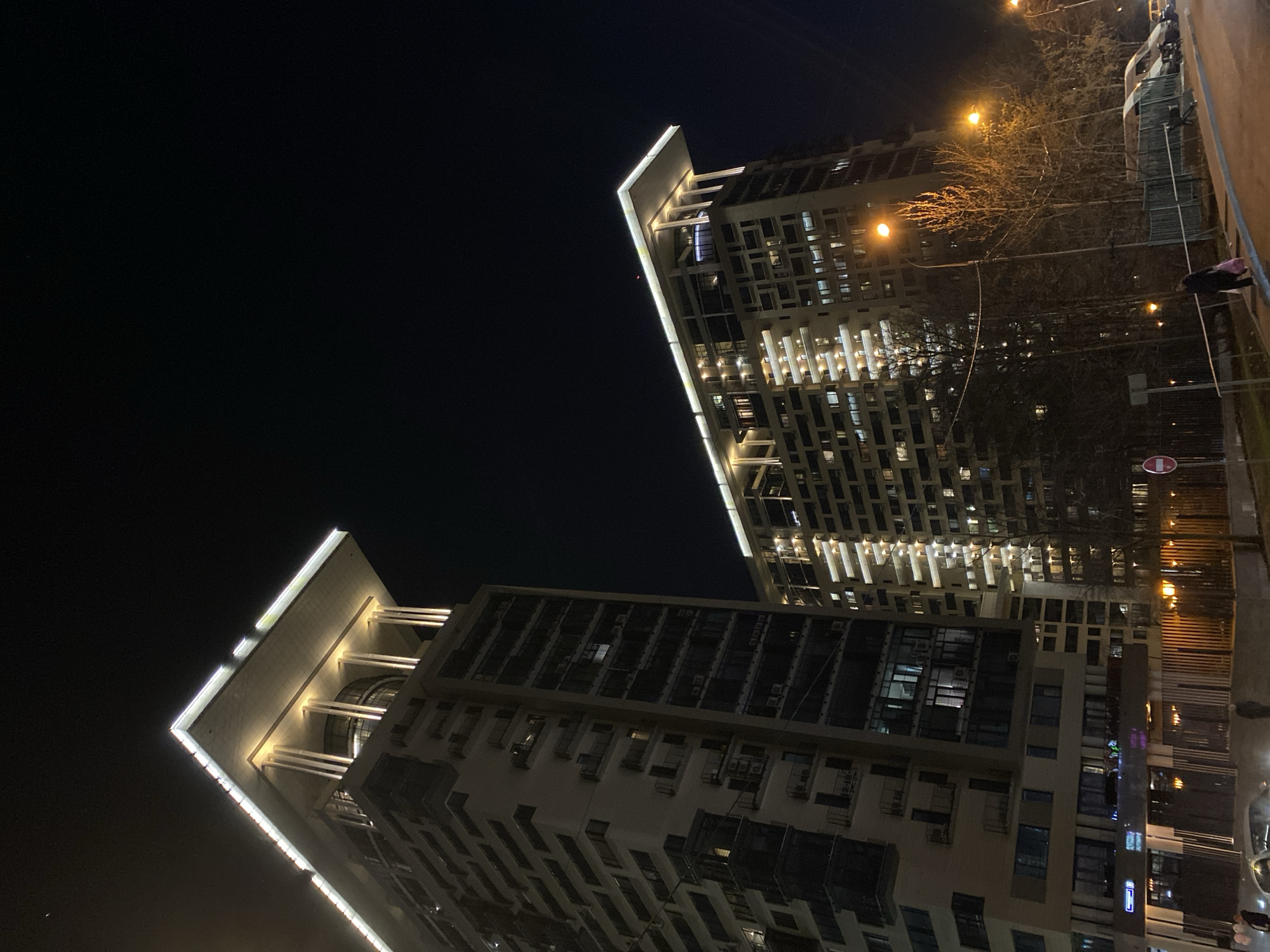 The image shows two tall buildings at night, likely in a densely populated urban area. The buildings are illuminated with lights on their facades, accentuating their architectural features. The sky is dark, indicating a night scene. On the right side of the image, a sidewalk runs along the edge of the frame, with sparse vegetation. The image appears to be taken at ground level, looking upwards towards the buildings. The presence of pedestrians suggests a public space. The buildings are likely residential or commercial. The location could be in Moscow, Russia.