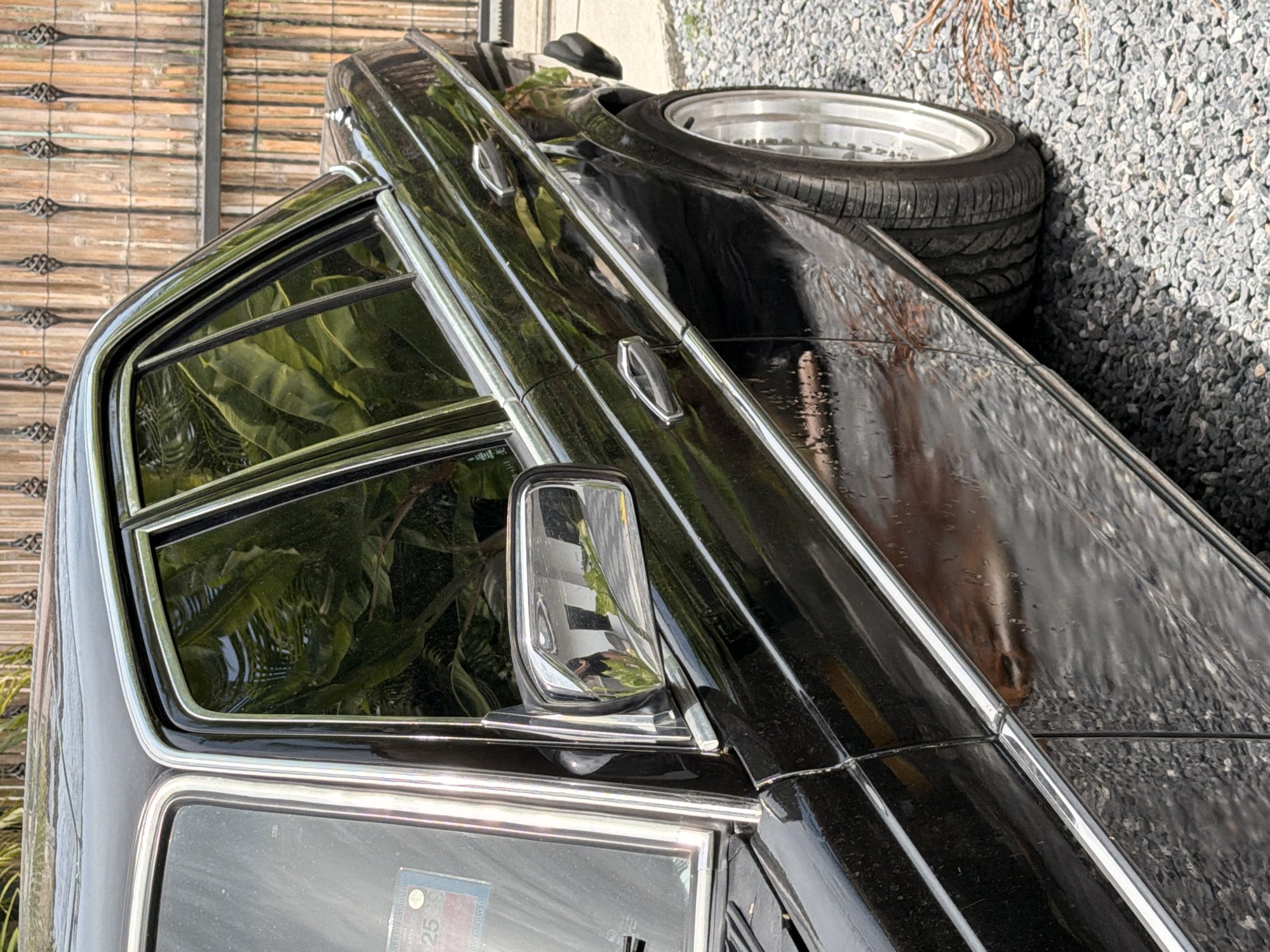 The image captures a close-up, angled view of the right side of a black Mercedes-Benz sedan parked on a gravel surface. The car's glossy black paint reflects the surrounding environment, including lush green foliage, a wooden fence, and a glimpse of what appears to be a white building in the side mirror, where a person is visible taking the photograph. The vehicle features chrome trim along the windows, doors, and a horizontal strip on the lower body. Two chrome door handles are visible. The rear wheel, with a chrome rim, is partially seen, with the tire resting on the gravel. In the background, a vertical wooden fence with decorative metalwork is prominent. A small sticker with the number "25" is adhered to the lower left corner of the driver's side window. The overall scene suggests an outdoor setting, likely in a residential or private area, possibly a driveway or a lot, given the gravel ground cover and the presence of a fence. The lighting indicates it is daytime. The location is identified as Ko Samui District, Thailand.