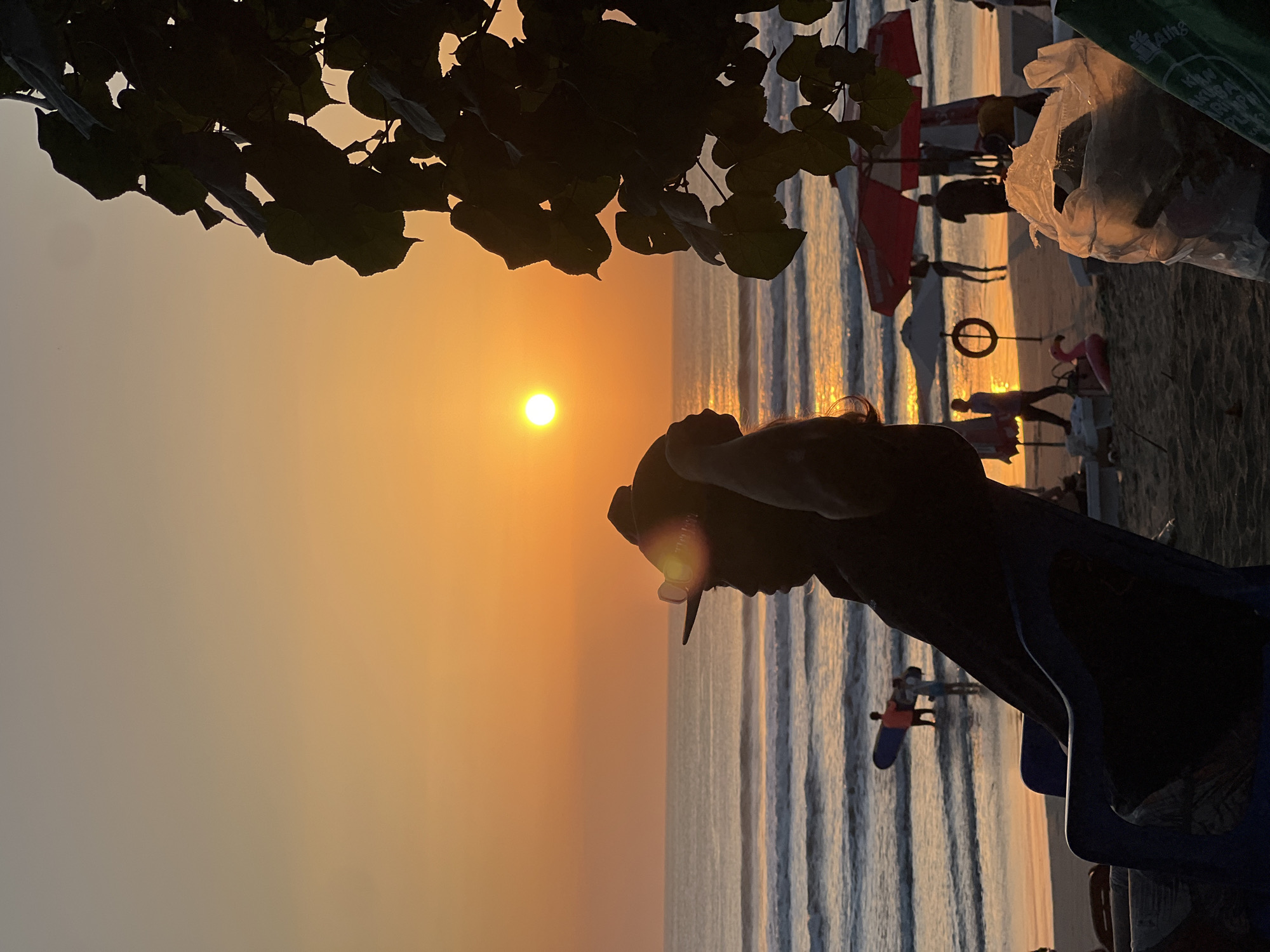 The image depicts a sunset on Kuta Beach, Indonesia. A person in silhouette is seated in a blue chair, facing the ocean and wearing a hat and sunglasses. The sun is setting over the water, casting a warm orange glow across the sky and sea. In the background, several people are visible on the beach and in the water. Some are carrying surfboards, while others are standing or walking along the shoreline. Beach umbrellas and a pink flamingo inflatable are also present. The foreground is occupied by the back of the chair and a plastic bag filled with trash. The overall scene conveys a peaceful and serene atmosphere.