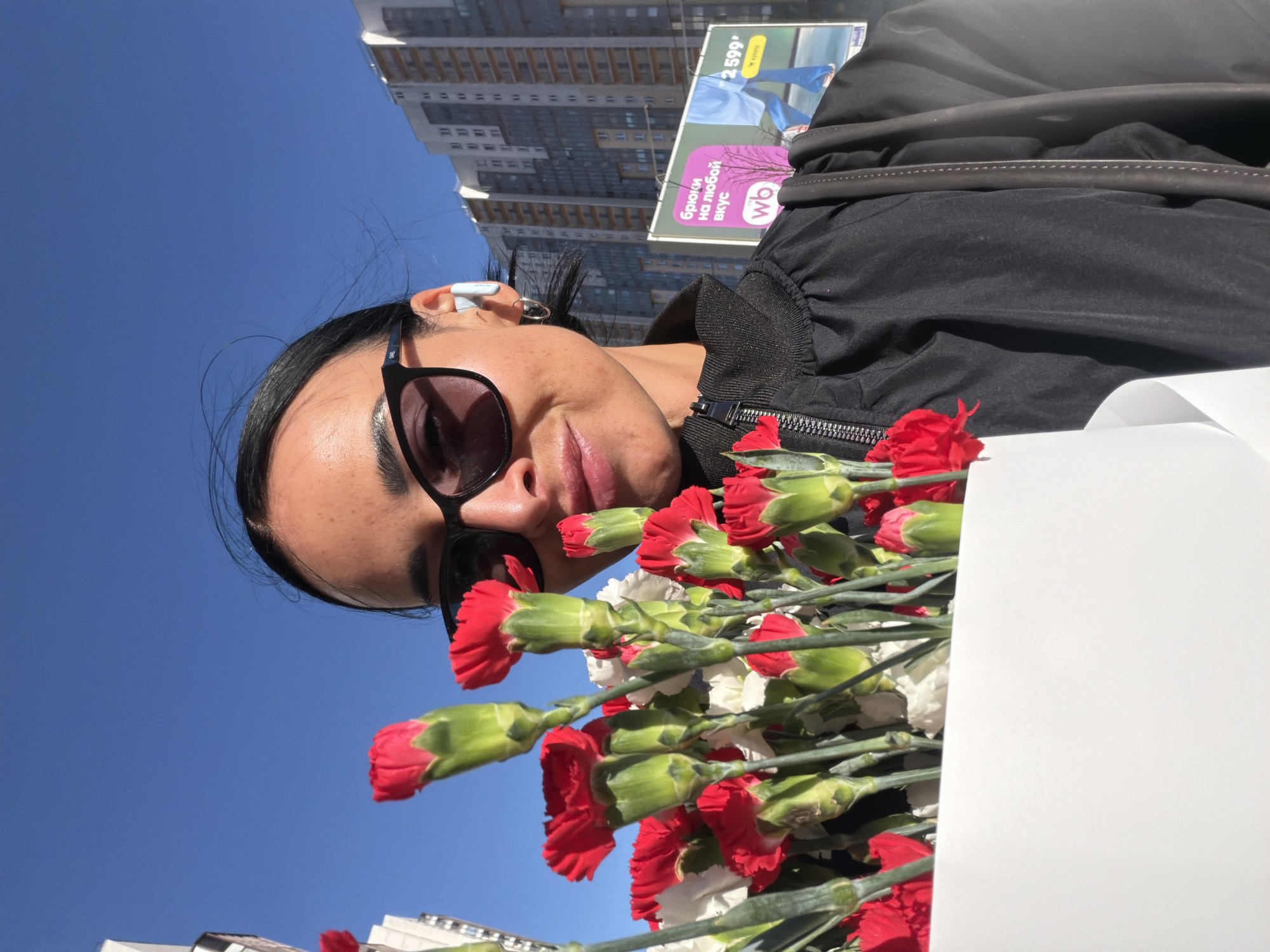 A woman with dark hair and sunglasses holds a bouquet of red and white carnations. She is wearing a black jacket and has a white earbud in her left ear. In the background, there is a clear blue sky and a tall building with numerous windows. A billboard is also visible, featuring advertisements in Russian. Based on the architectural style and presence of Russian signage, the location is likely Saint Petersburg, Russia.