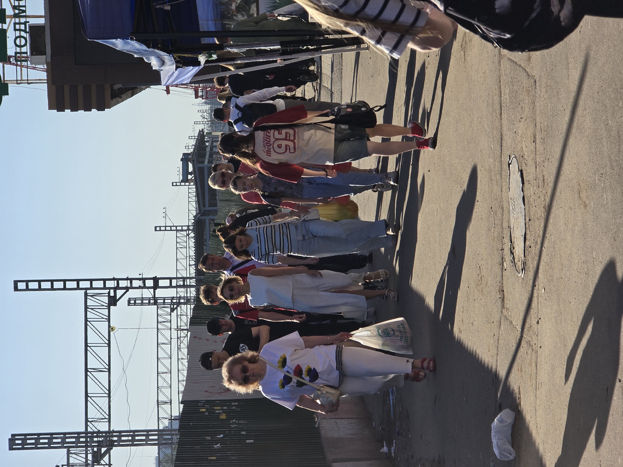 A ground-level, horizontally-oriented shot captures a queue of approximately ten people. The subjects are primarily standing on a weathered, concrete pavement adjacent to what appears to be a railway or tram structure to the left.
A woman in the foreground wears a white t-shirt with floral graphics and white pants, carrying a shopping bag and a small purse. Moving backward, diverse individuals make up the line, some in casual clothing, others in more formal attire. Some are wearing caps and carrying bags.
The background includes a partially constructed building or scaffolding structure made of metal beams and pillars. The weather appears sunny, as suggested by the crisp shadows cast by the subjects and nearby objects. There are signs of urban decay, such as debris and wear on the pavement. The location is identified as Khimki, Russia.
