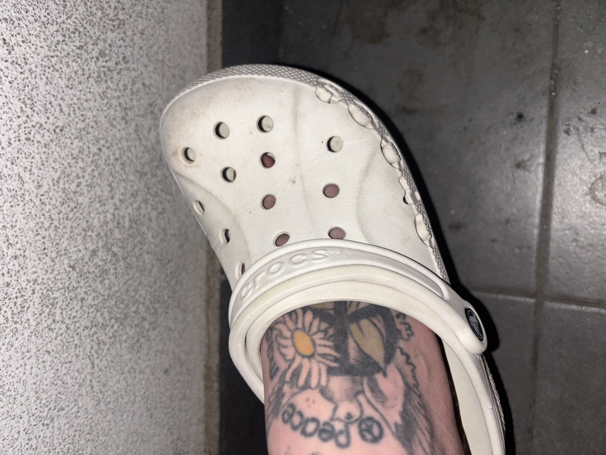 A close-up photograph depicts a person's foot adorned with a tattoo featuring a daisy, the word "peace," and additional floral and symbolic elements. The foot is clad in a white Croc sandal. The backdrop includes a textured wall on the left and tiled flooring on the right. There is no contextual information to indicate the location is Belgrade, Serbia.
