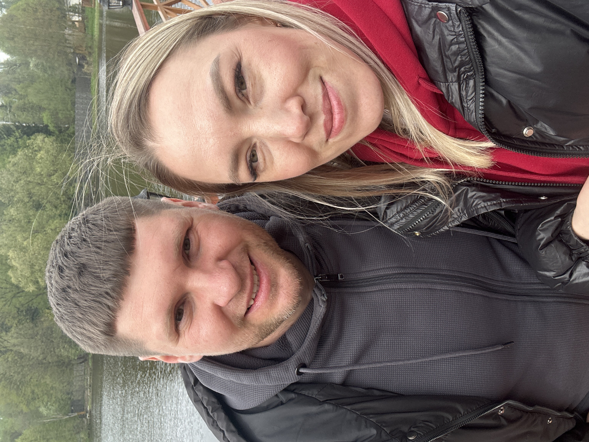 A selfie depicts a man and a woman outside near a body of water, possibly a lake. The woman, positioned at the top of the frame, has blonde hair and is wearing a red hooded sweatshirt under a black puffer jacket. The man, located below the woman, has short, dark hair and is wearing a gray hooded sweatshirt and a black jacket. The background features a body of water with trees on the opposite bank. The location is likely Grebnevo, Russia.