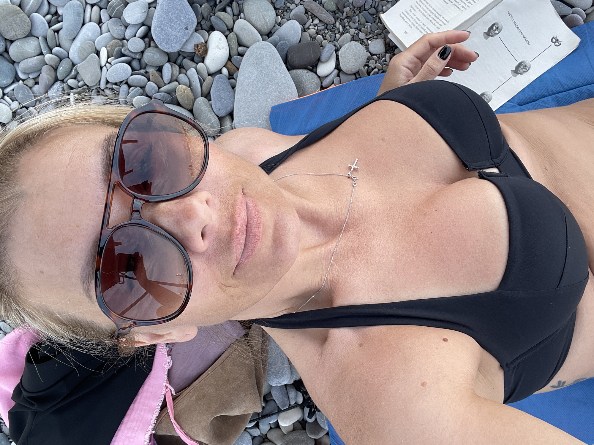 A woman is lying on a blue mat on a pebbled beach, with a book in her hand. She is wearing a black bikini and sunglasses. The book appears to be a novel, with Russian text visible on the open page. The woman's hand, with black nail polish, is holding the book. The sun is shining, casting shadows on the pebbles and the woman's skin. The scene appears to be a peaceful moment of relaxation. The visible text in the image is in Russian and includes the title "ЧАСТЬ 1. МАСКИ КАРАКАЛЛЫ," which translates to "PART 1. MASKS OF THE CARACALLA." Other visible text includes "Меня звали Варий Алет. Манго бага," and "Загавал."