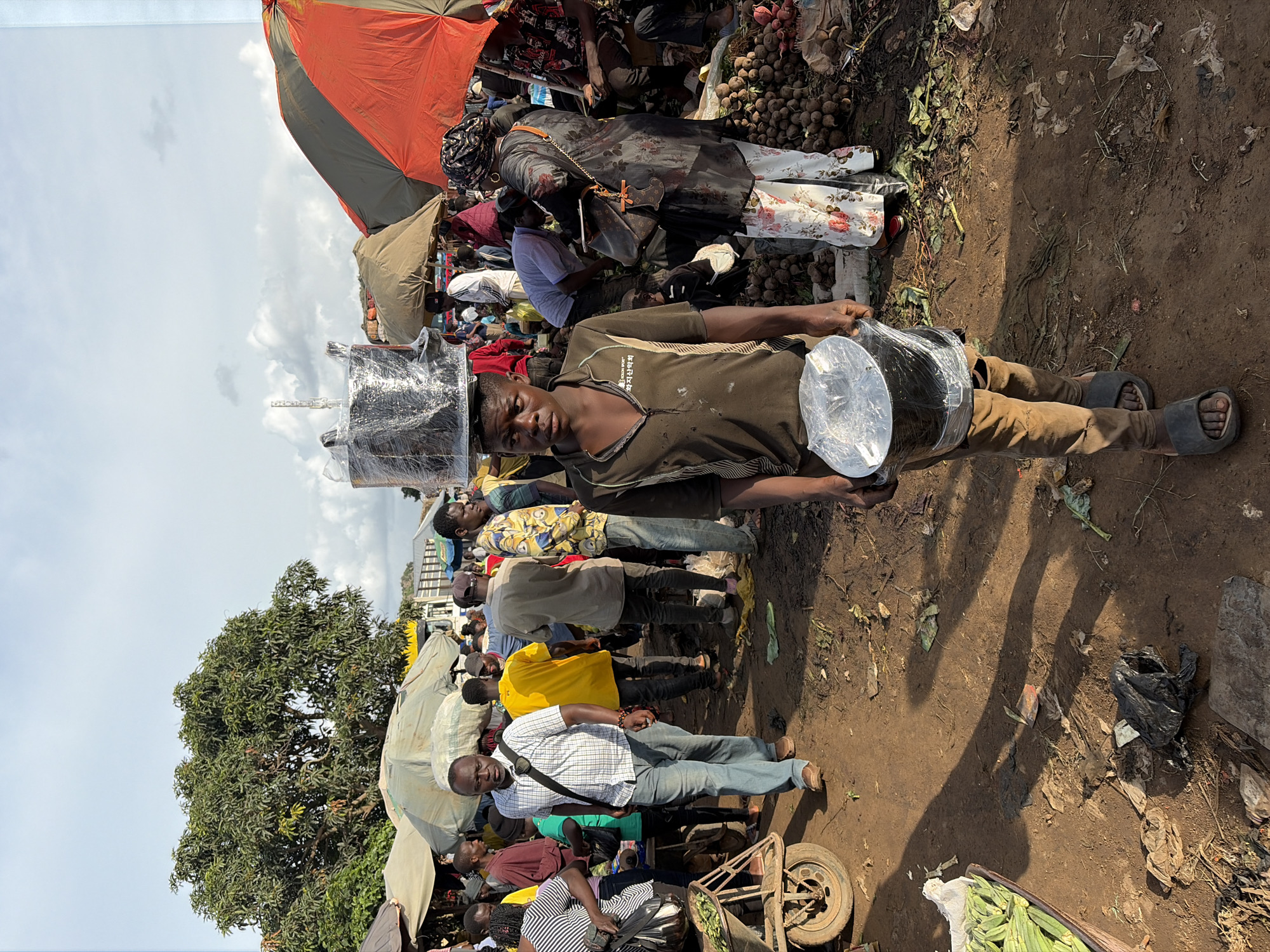 In Gura Top, Nigeria, a young Black male walks through a bustling outdoor market, balancing a large, circular object wrapped in clear plastic on his head. He is wearing a short-sleeved, dark brown polo shirt with writing in an East Asian language on the left side of his chest, and light brown trousers. In his hands, he carries a smaller, similarly wrapped circular object.
The market is crowded with people browsing and selling goods. Various stalls are visible, some covered by umbrellas of orange, green, and tan. Piles of produce, appearing to be root vegetables like beets or potatoes, are stacked on the ground in the background. The ground is dirt and debris-strewn. Other individuals are seen walking, carrying items, and interacting with vendors. A man in a checkered shirt and jeans walks past in the foreground to the left, and other shoppers are visible further back. The sky above is partly cloudy.