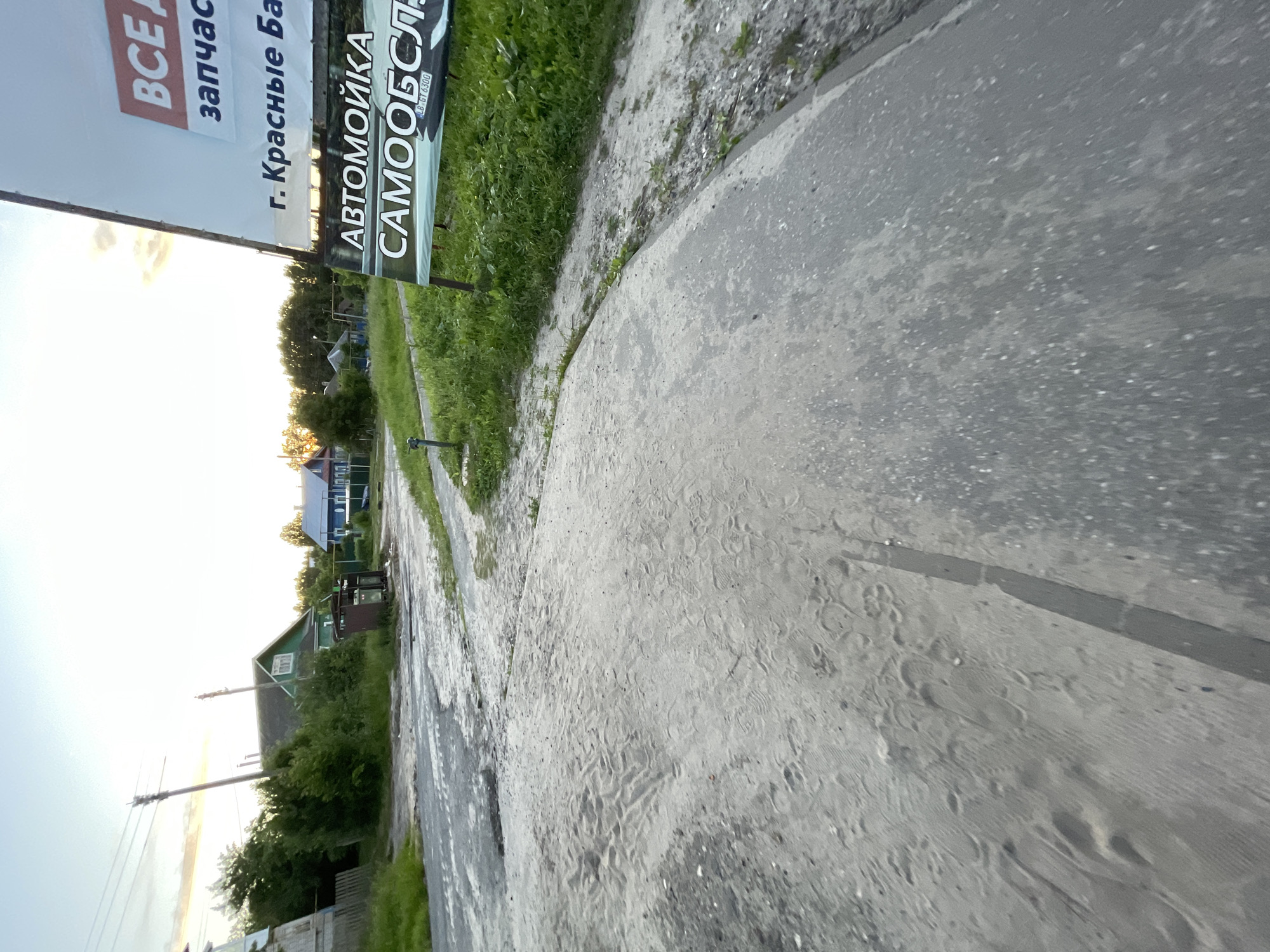 The image depicts a rural scene in the village of Krasnye Baki, Russia, during what appears to be sunset. In the foreground, a sandy, uneven path leads into the frame, with tire tracks visible on the surface. To the right, a large billboard advertises "ВCE A" (possibly "Все Авто" - All Cars) and "запчасти" (spare parts), along with "г. Красные Баки" (Krasnye Baki village). Below this, it advertises "АВТОМОЙКА САМООБСЛУЖИВАНИЯ" (Self-service car wash). The background shows a few houses, including a green-roofed building, and lush green trees. Telephone poles and power lines are also visible, indicating infrastructure in the area. The sky is a pale orange and yellow, characteristic of sunset. There are no people or animals present in the image.