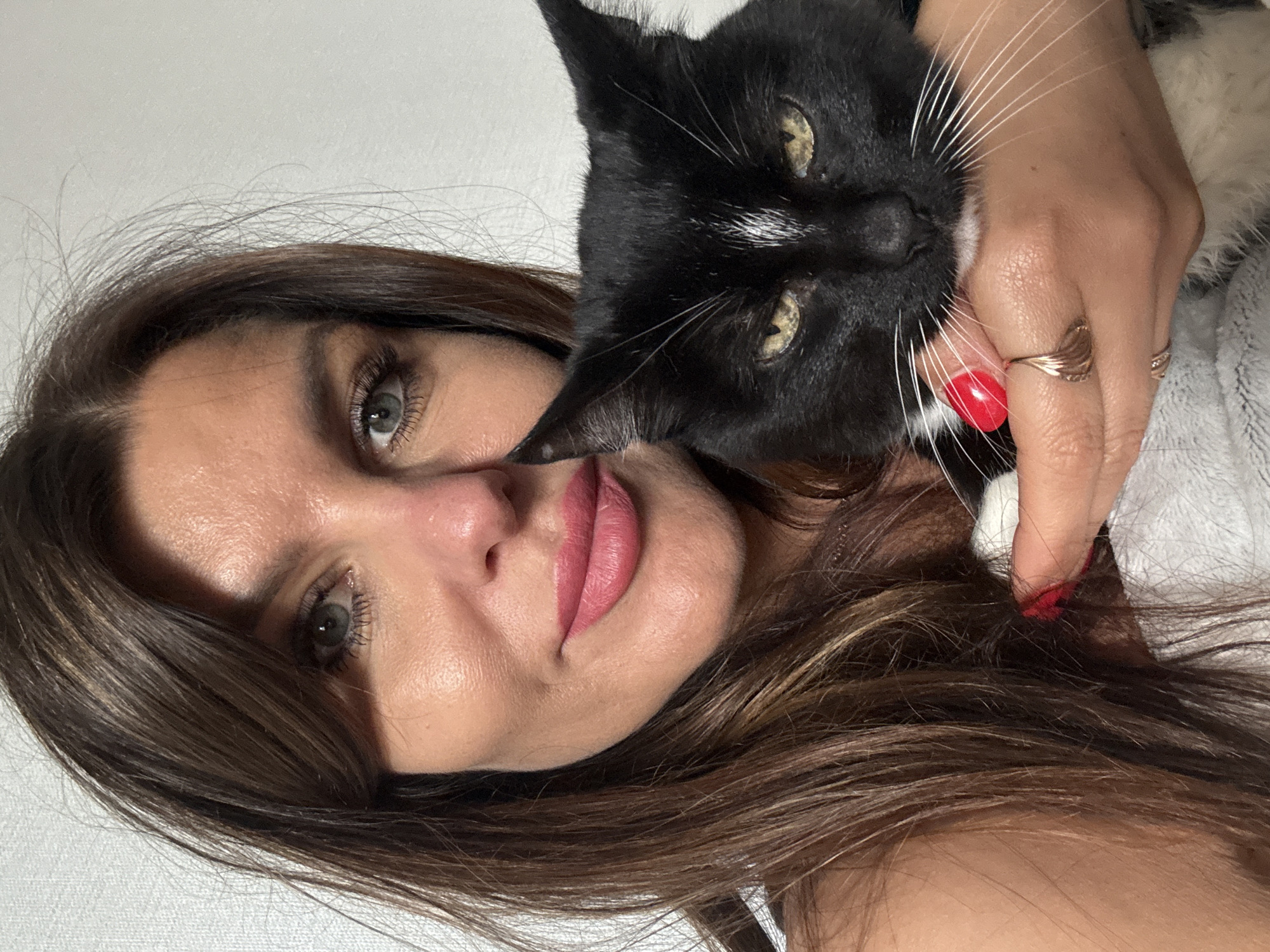 The image displays a close-up indoor portrait of a woman and a black and white cat. The woman, positioned on the left, has long, dark brown hair with lighter brown highlights, parted on the left side, and some strands framing her forehead. Her green eyes are open, and she has pink lipstick on her lips, with a slight, gentle smile. Her skin texture is visible, with some pores.
She is holding a black and white tuxedo cat with her right hand, which is visible in the lower right portion of the image. Her fingernails are painted bright red, and she wears a gold-toned ring with a textured, curved design on her right ring finger. The cat has predominantly black fur, prominent white whiskers, a white patch on its chest/chin, and a distinctive thin white vertical stripe between its eyes. Its eyes are yellow-green and appear somewhat narrowed or sleepy. The cat's head is angled slightly towards the right, and its gaze is directed generally forward.
The background is a plain, light-colored (likely white or off-white) wall with a subtle, rough texture. There are no other discernible objects or specific environmental features in the background. The image does not contain any visual identifiers to confirm its location as the city of Chaikovsky, Russia.
