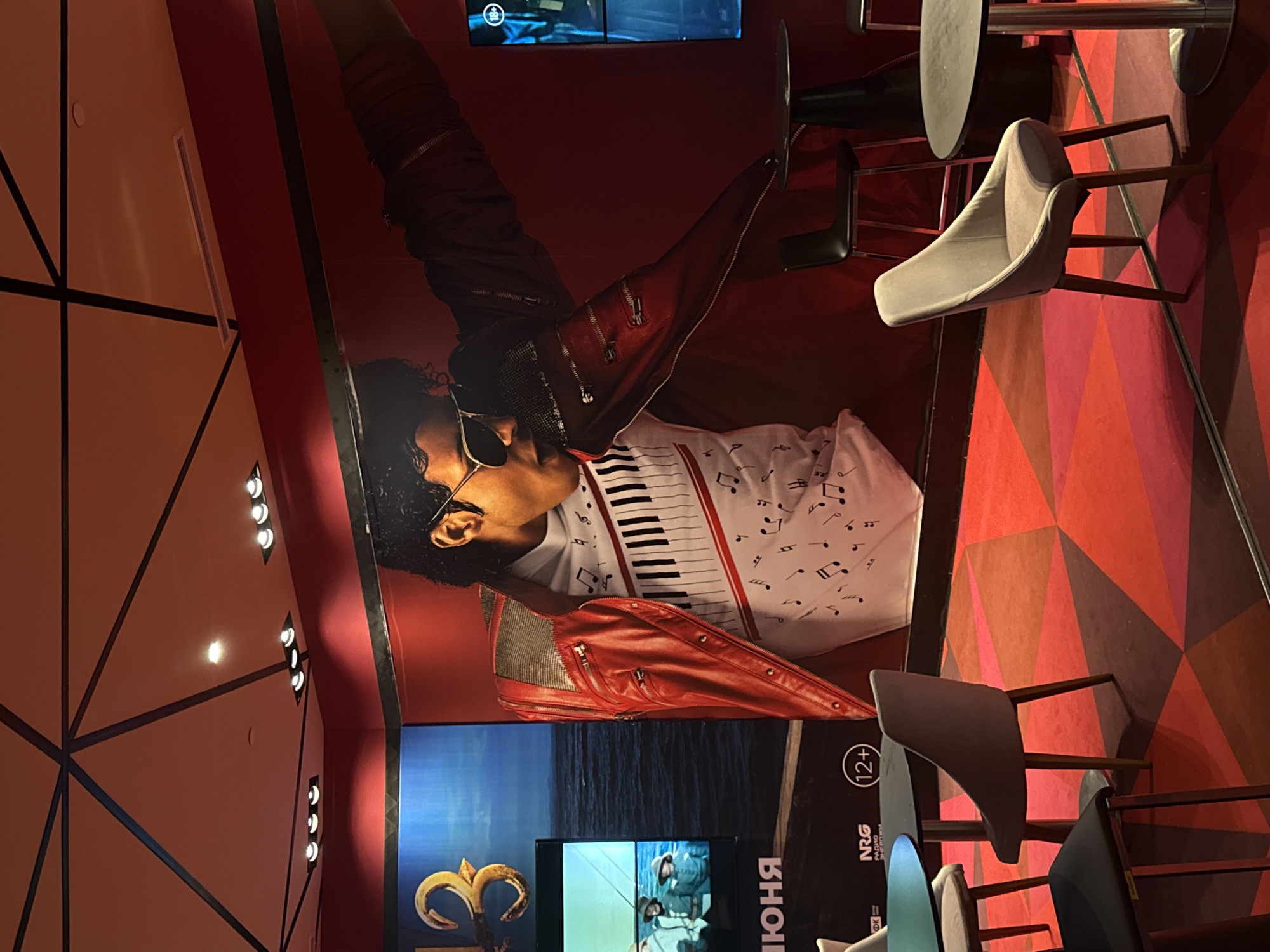 This image depicts a room with a large mural of Michael Jackson on the wall. The mural shows him wearing a red leather jacket and a white t-shirt with a piano key design and musical notes. In the background, there is a movie poster featuring the number "3" and a scene with people on a boat.
The room has a red carpet with a geometric pattern. There are several tables and chairs, including black chairs at round tables and lighter-colored chairs positioned around other tables. One of the chairs is a light gray upholstered chair with wooden legs. Another chair is dark gray and appears to be a stool.
On the wall to the left of the Michael Jackson mural, there is a screen displaying the movie poster. To the right of the mural, another screen is partially visible, showing content with an "18+" rating.
The ceiling features a geometric design with dark lines and recessed lighting. The overall ambiance suggests a waiting area or lounge, possibly within a cinema or entertainment venue. The poster on the left has text in Russian that includes "ИЮНЯ," indicating June.
The location is identified as Khimki, Russia.