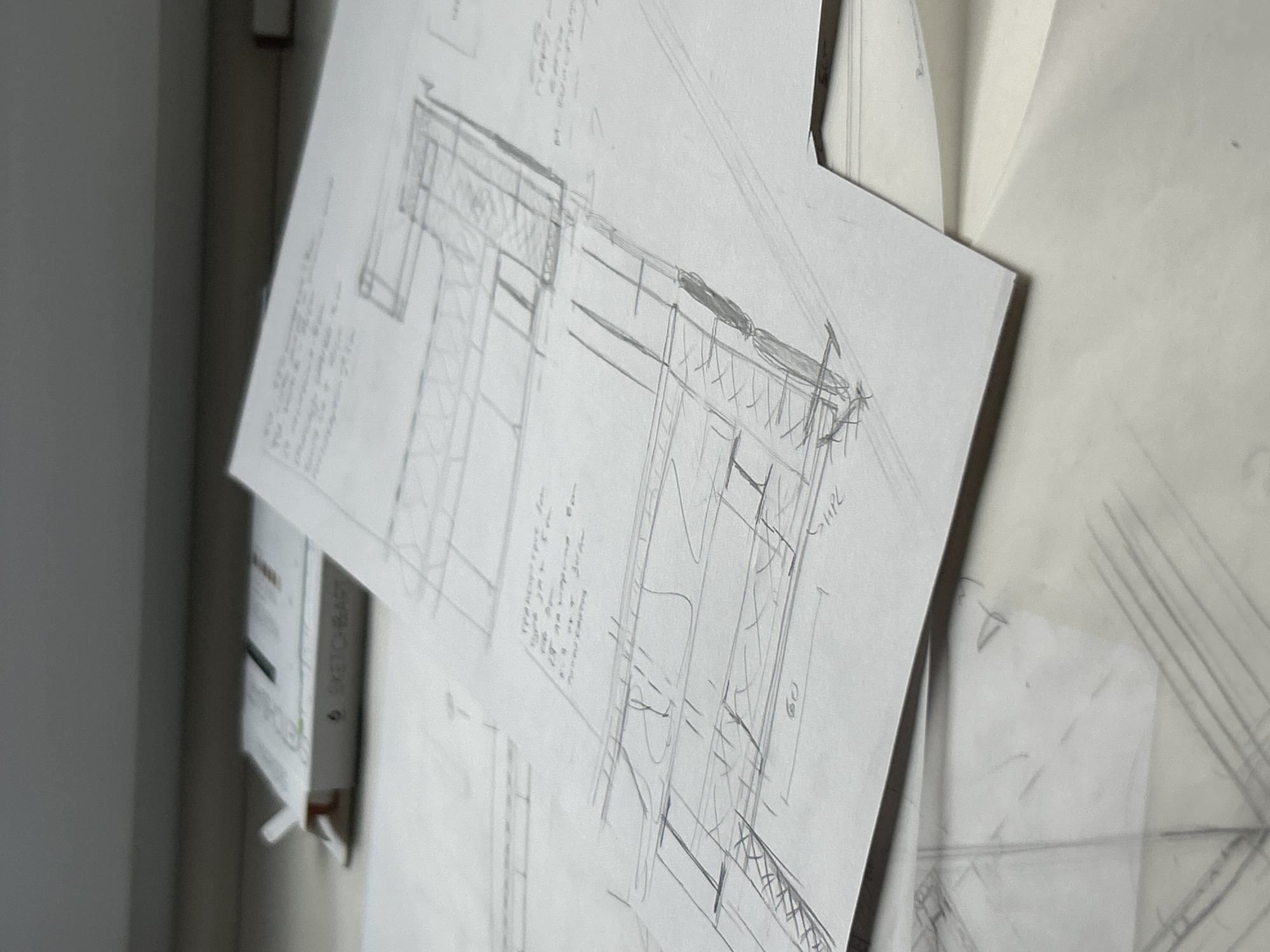 The image shows architectural sketches attached to a wall, likely in an office or studio. The sketches appear to be pencil drawings on white paper, possibly depicting building details or sections, with notes in a hand-written style. The paper is attached to the wall with pins and/or adhesive tape. The background is blurred, but a white wall is visible. A vertical object to the left of the sketches appears to be a box or container with the word "SKETCH-BAR" visible on it. The location is not discernible from the image.