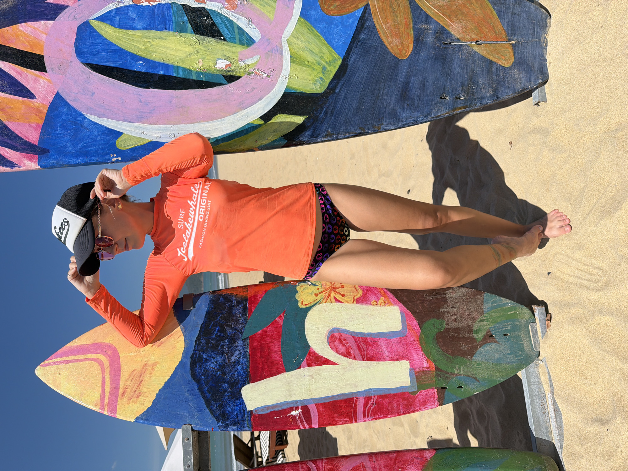 A woman stands on a sandy beach in Quy Nhon, Vietnam, between two vibrantly painted surfboards. She is wearing an orange long-sleeved rash guard with "SURF LakeWhale ORIGINAL FASHION DIVING SUIT" printed on it, and black bikini bottoms with a colorful geometric pattern. A black and white trucker hat with sunglasses is perched on her head, and she is adjusting the brim with her left hand. The surfboard on her left is painted with abstract shapes in shades of yellow, pink, blue, and red, featuring a large white "h". The surfboard on her right has a blue background with large, stylized pink and green abstract shapes and orange floral elements. The sun is shining brightly, casting shadows on the sand. In the background, the ocean and a clear blue sky are visible, along with beach chairs and umbrellas.
