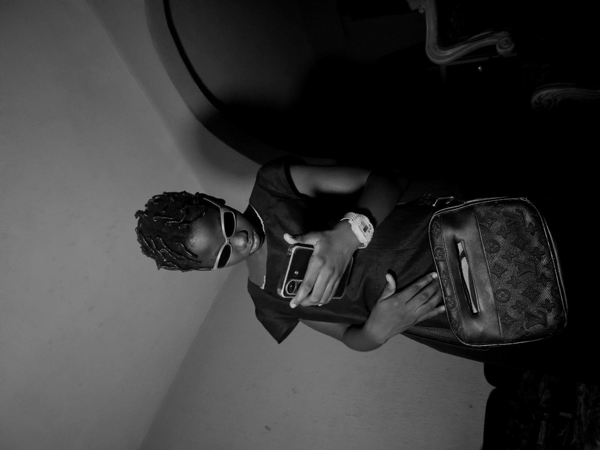 A black-and-white photograph shows a person indoors, possibly in a room or a building in Nkwelle, Nigeria, as specified. The person is wearing sunglasses and a watch on their wrist, holding a smartphone. They have a bag with a pattern on it resting on their side. The background is partially obscured, with a wall visible on one side and a dark area that may contain furniture. The photograph appears to be taken at an unusual angle, with the person seemingly tilted or the camera positioned at an angle.