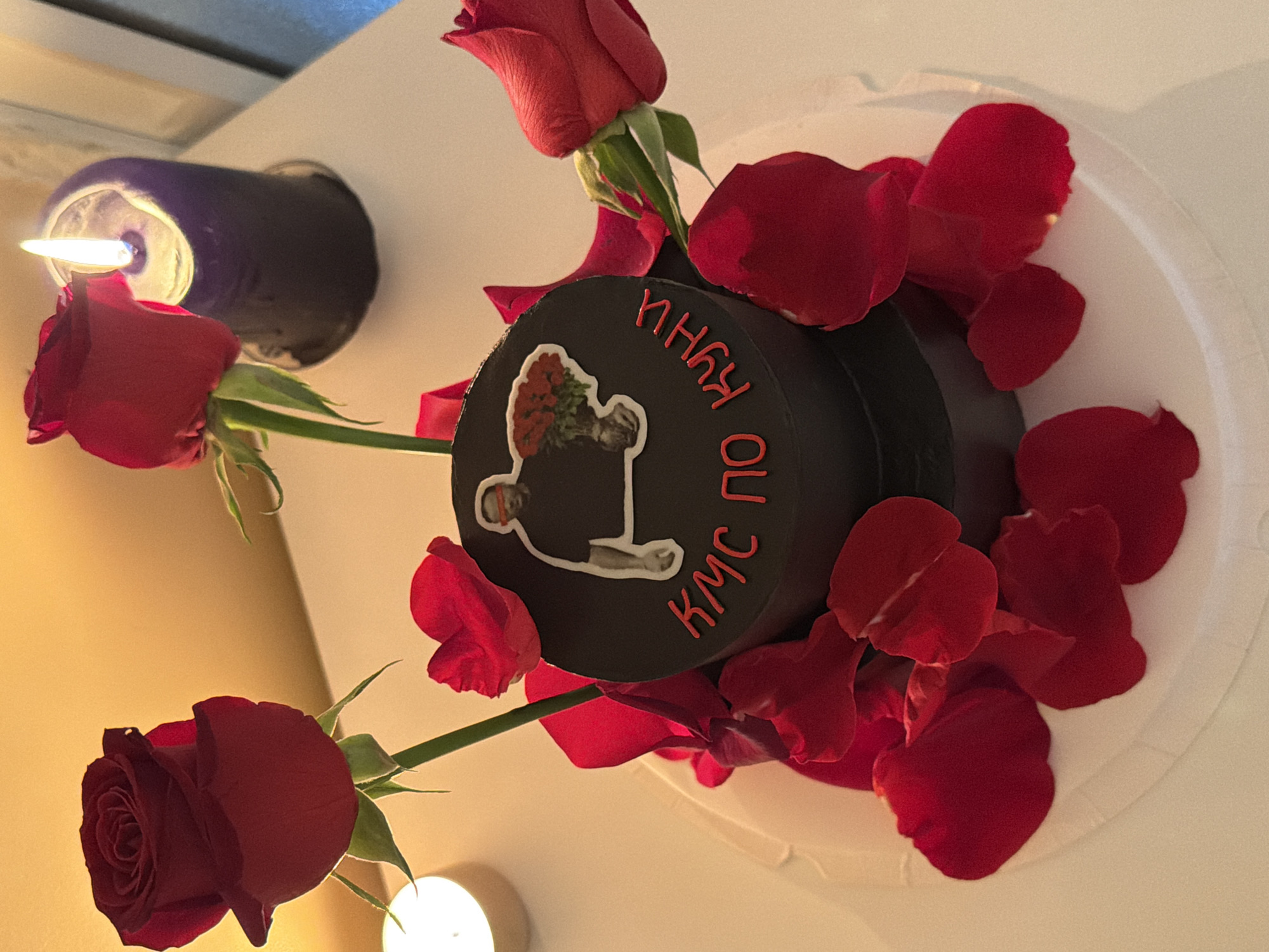 A top-down shot shows a black, circular cake decorated with red rose petals and three red roses with green stems. A sticker of a man holding flowers is on the cake's surface, and Cyrillic lettering surrounds it. A lit candle in a dark holder is to the left of the cake. The cake sits on a white plate. The image suggests an indoor setting. There is no indication of the location as Tel-Aviv, Israel.