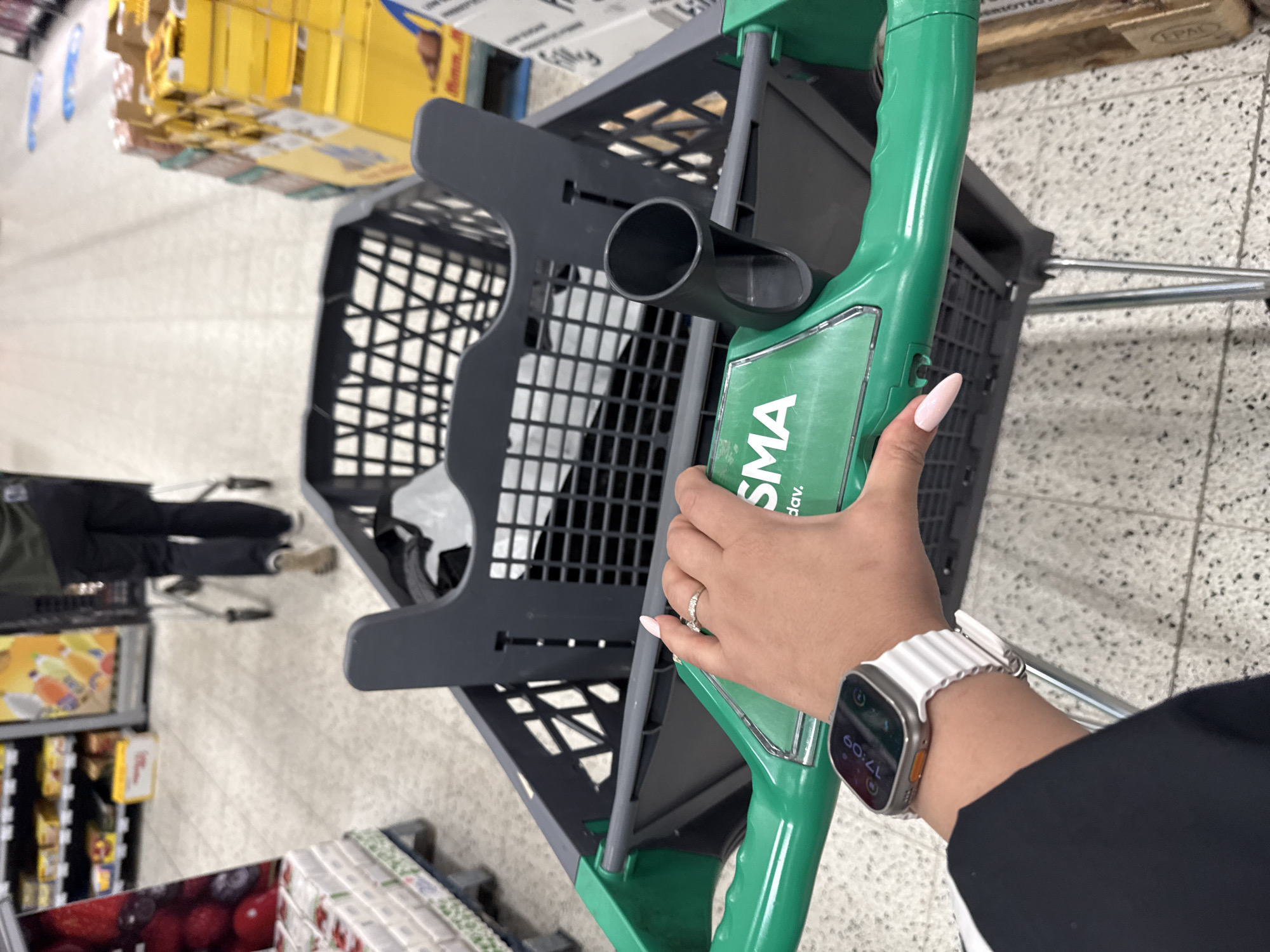 A first-person perspective shot shows a hand with a white Apple Watch and light pink acrylic nails gripping the green handle of a shopping cart in a supermarket aisle. The shopping cart is mostly empty, with a black plastic basket visible. Behind the cart, other shoppers and shelves stocked with various products are out of focus. The floor is tiled with a light gray and black speckled pattern. The environment suggests a retail setting with bright, artificial lighting. The word "SMA" is visible on the green handle of the shopping cart. The image does not contain enough information to determine the specific location within Tallinn, Estonia.