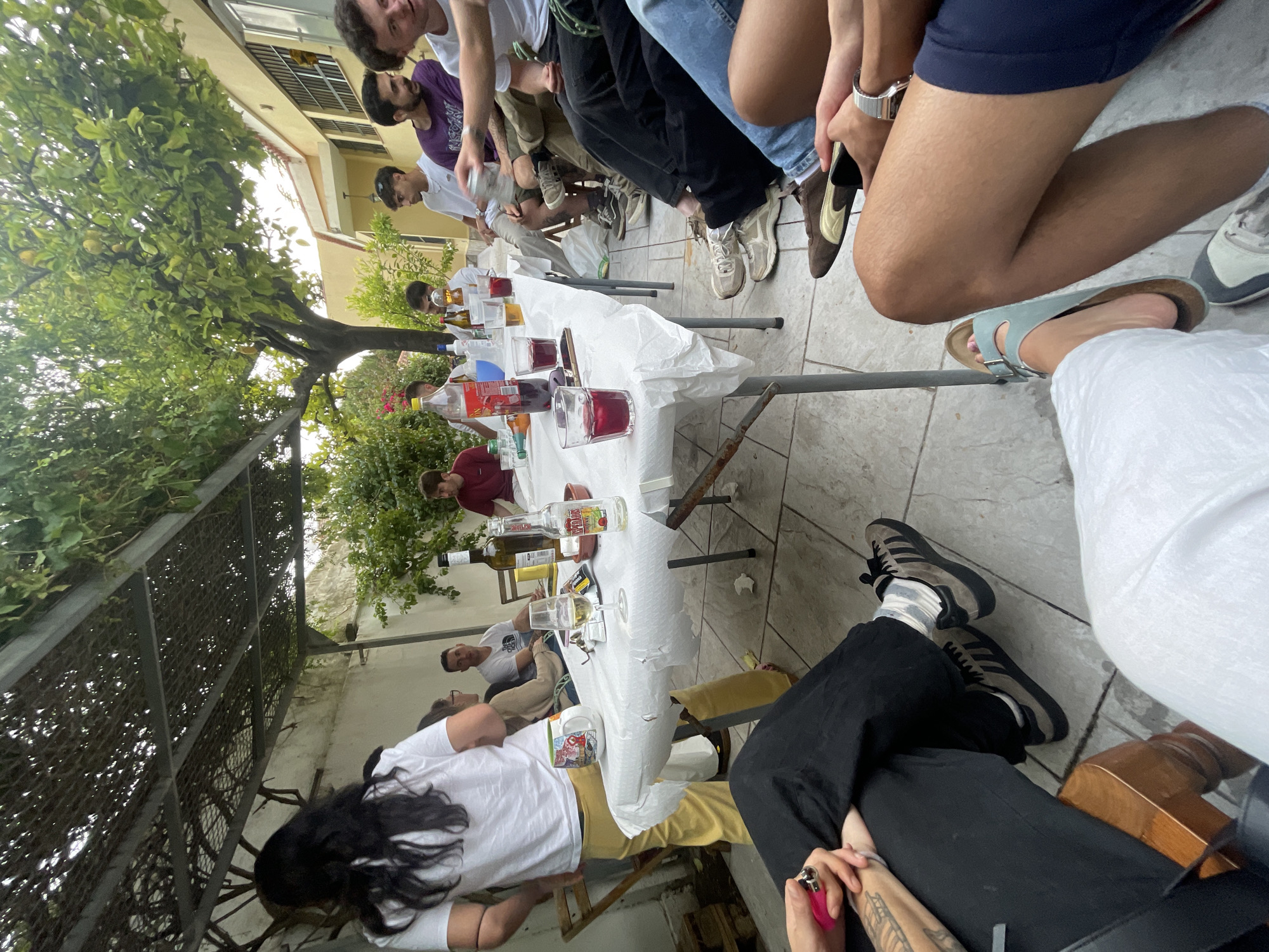 A group of people are gathered around a table outdoors. The table is covered with a white cloth and has various beverages and glasses on it. The people are dressed casually and appear to be socializing. The location appears to be a courtyard or patio in Jerez De La Frontera, Spain, with a tree providing shade and buildings visible in the background. There are at least 12 people gathered around the table with more potentially out of frame.