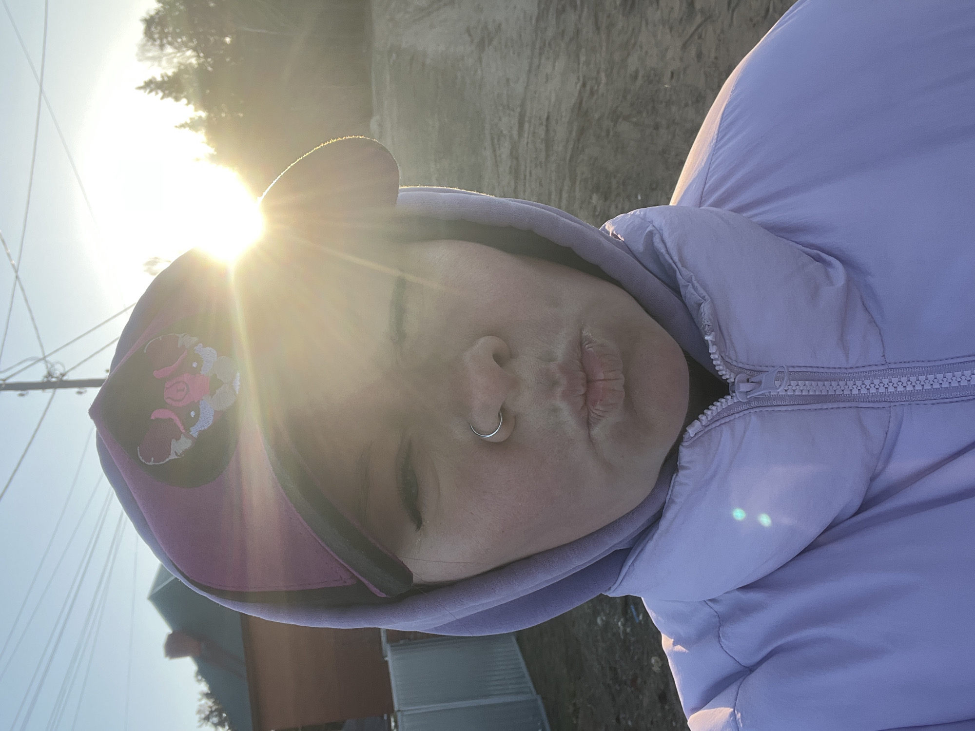 A close-up photograph shows a person wearing a light purple hooded jacket and a cap with a cartoon image on it. The person has a nose ring and is making a pouting expression. The image is brightly lit with sunlight, causing glare. The background includes trees, power lines, and buildings, suggesting an outdoor environment. Based on the EXIF data, the image was taken in the city of Chaikovsky, Russia.