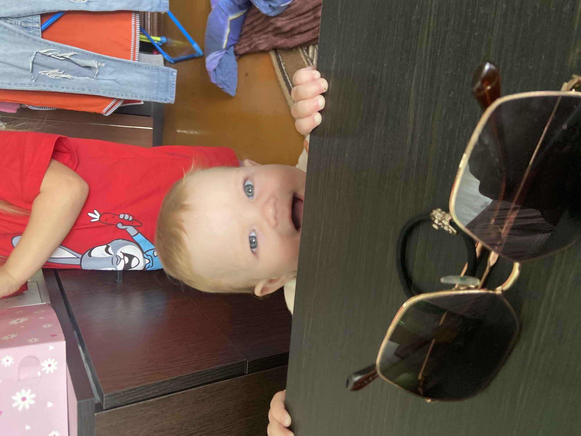A baby with blond hair and blue eyes, wearing a red shirt with a cartoon character on it, is peering out from behind a dark-colored wooden surface. The baby's mouth is open, and their hand is visible gripping the edge of the surface. A pair of sunglasses with gold frames and dark lenses is hanging on the edge of the same surface. Other objects, including clothes and a pink box with white daisy patterns, are visible in the background. The specific location in Фоки, Russia, is not discernible from the image.