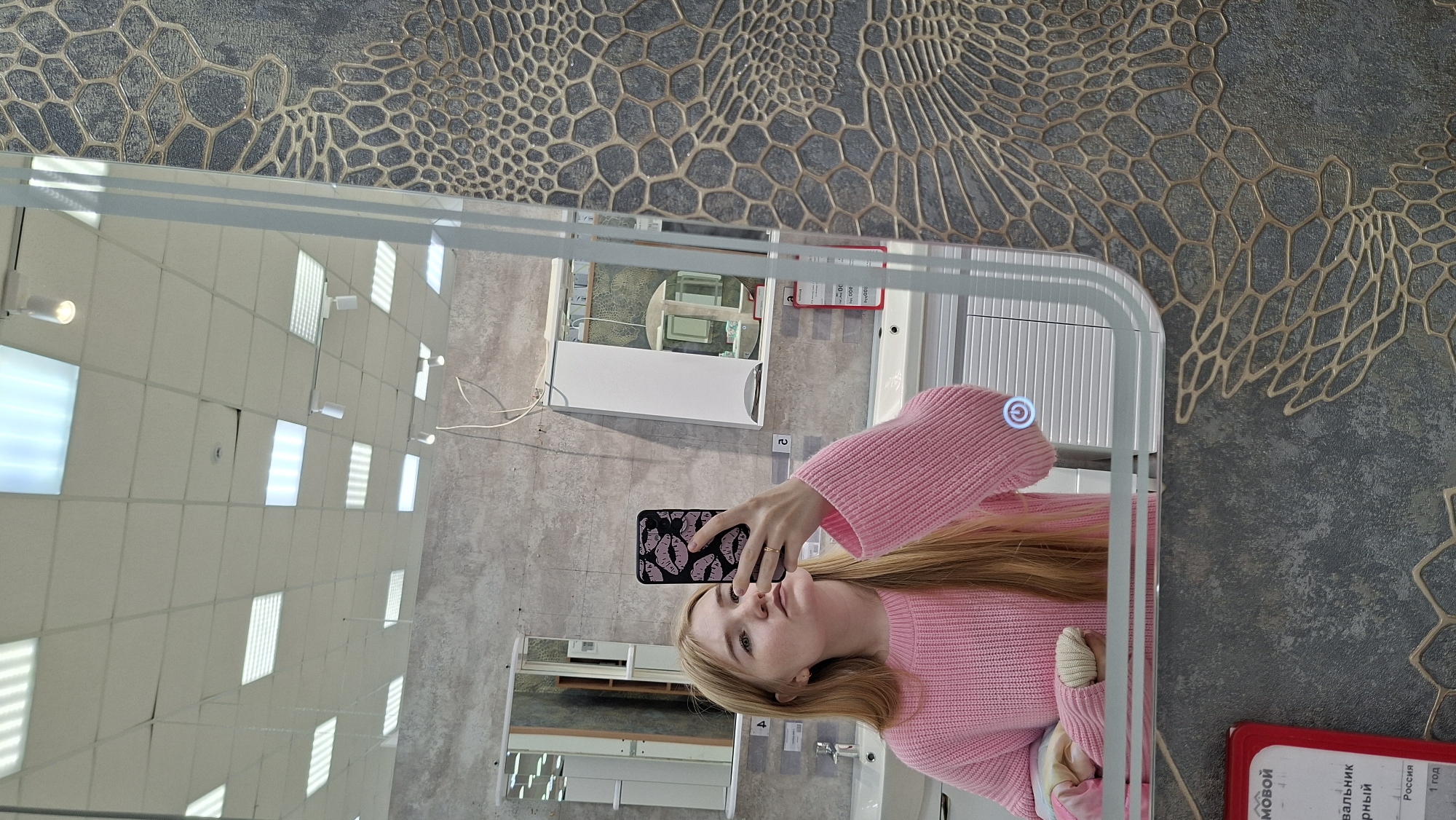 The image depicts a woman with long blonde hair, wearing a pink sweater, taking a selfie in a mirror. The mirror is framed and has an integrated LED light. She is holding a black smartphone with a repeating pattern of lips on the case. The background reflected in the mirror shows a showroom or retail space, possibly for bathroom fixtures, as evidenced by the display of various sinks and mirrors. The ceiling is tiled with fluorescent lighting panels. The foreground around the mirror shows a wall with a decorative pattern of gold hexagonal outlines on a gray background. A red sign with Russian text is partially visible on the lower right of the image.
It can be inferred from the sign, which indicates the item is a "sink" and "made in Russia," that this photograph was taken in a store in Russia that sells bathroom fixtures. Further geographic information is conveyed by the store's location in Chaikovsky, Russia.