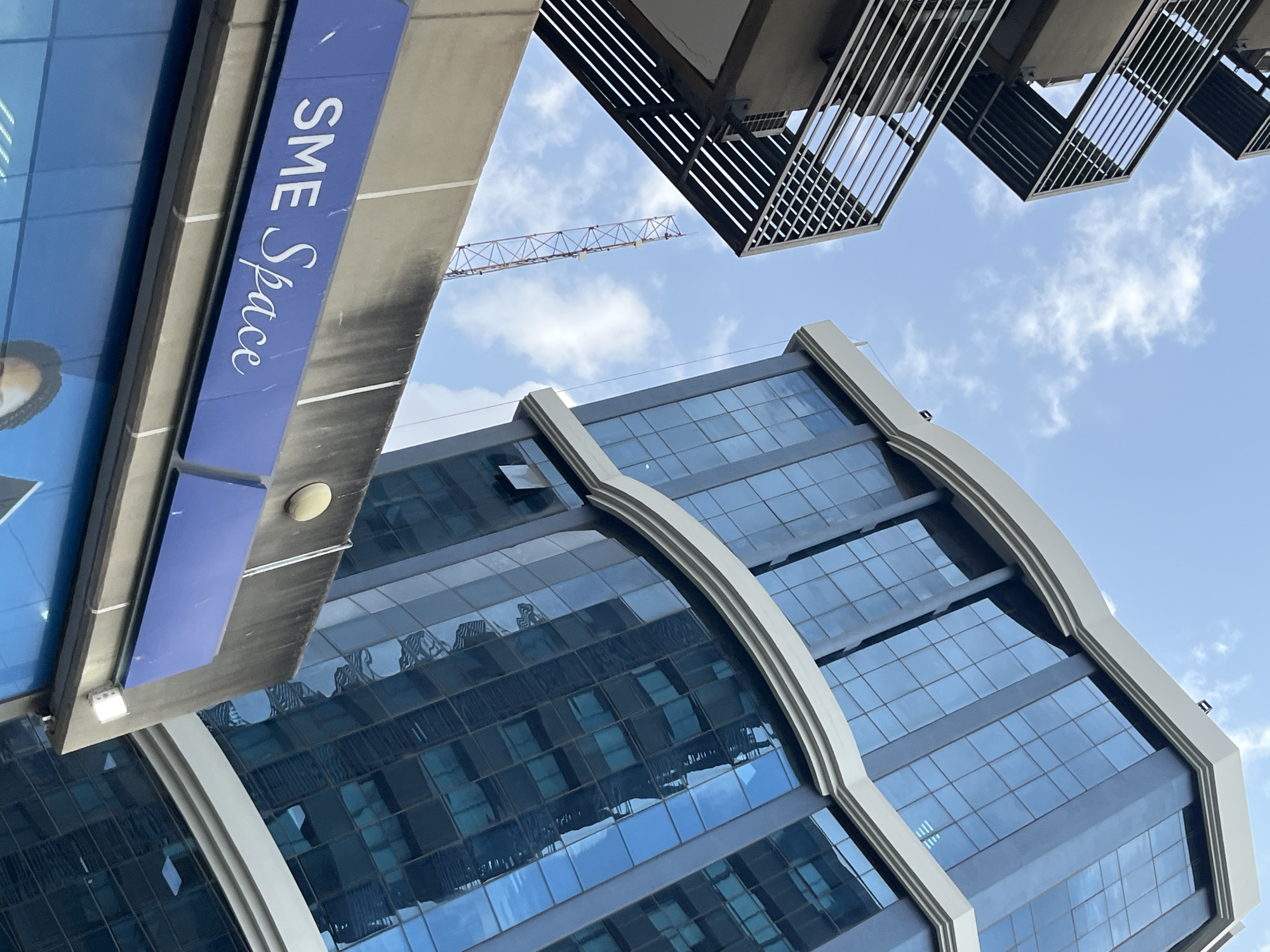 The image shows a low-angle view of the "SME Space" building located in Dar es Salaam, Tanzania. The building features a modern architectural design with a facade of blue glass windows and a white curved trim. Adjacent to it, an external metal staircase is visible, extending upwards against a clear blue sky with scattered white clouds. In the background, a tall communication tower stands against the sky. The lower portion of the image reveals a blue signage with the text "SME Space" in white lettering. The overall scene depicts an urban environment with commercial buildings and infrastructure.