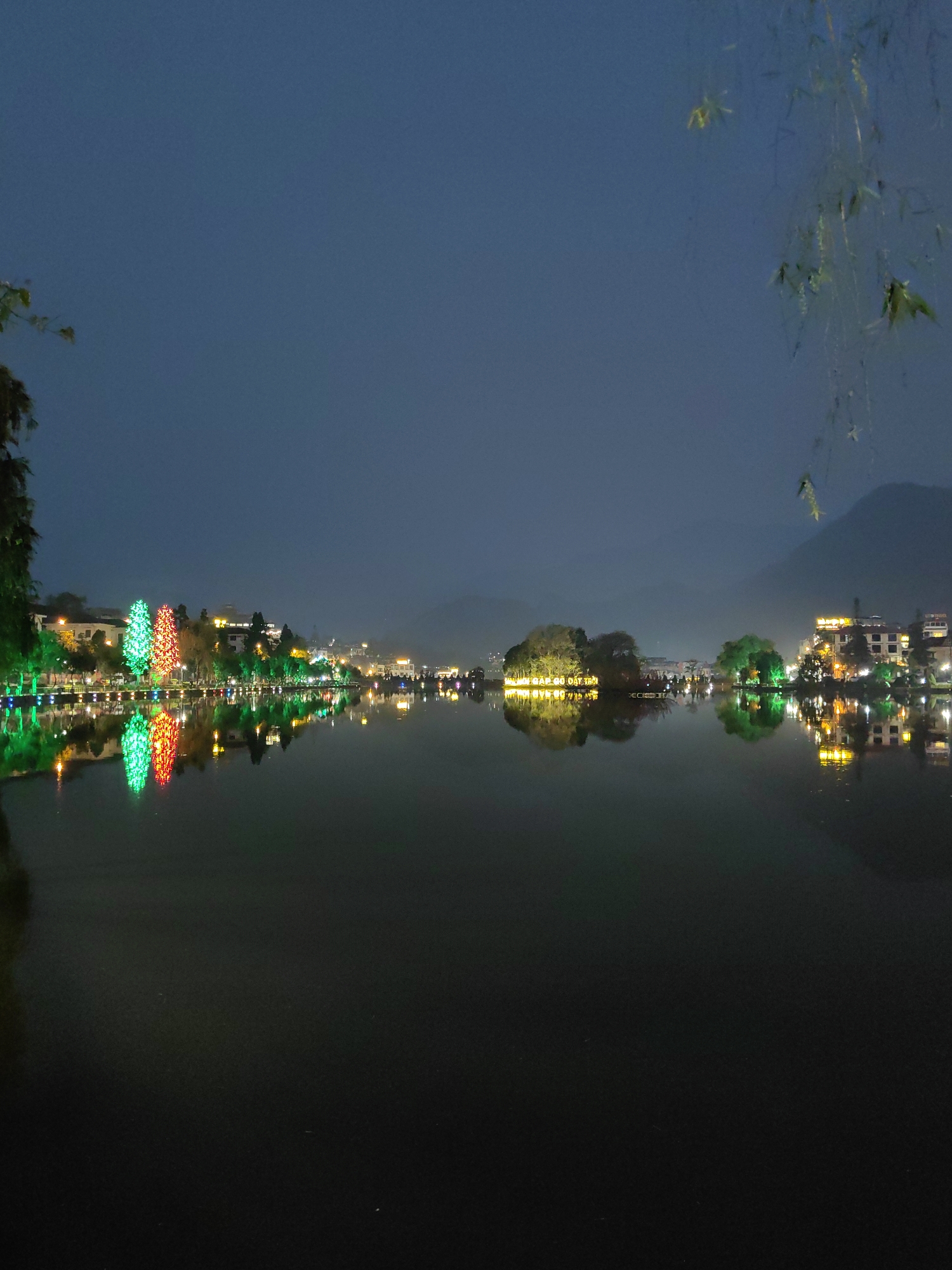 The image depicts a nighttime panoramic view of Sa Pa Lake (Hồ Sa Pa) in Sa Pa, Lào Cai Province, Vietnam. The scene is dominated by the still, dark water of the lake, which occupies the lower two-thirds of the frame, perfectly reflecting the artificial lights from the surrounding town and shoreline.
Along the left shoreline, a sequence of illuminated buildings and numerous trees adorned with various colored lights are visible. Prominently, two large, cone-shaped trees, one illuminated bright green and the other bright red/orange, stand out. These lights, along with other streetlights and building illuminations, create distinct, inverted reflections on the lake's surface.
Towards the center-right of the lake, a small landmass or island features illuminated trees and what appears to be a structure with glowing text, which also reflects on the water. The right shoreline shows additional buildings and foliage, brightly lit, with their reflections mirrored below.
The sky, occupying the upper third of the image, is a dark, hazy blue-gray, indicative of night. Distant mountains or hills are faintly discernible in the far background on the right, partially obscured by a light atmospheric haze. Portions of tree branches with sparse leaves are visible in the extreme upper left and upper right corners, framing the scene. No individuals or specific actions are visible. The overall environment is urbanized around a natural lake, illuminated for aesthetic purposes at night.