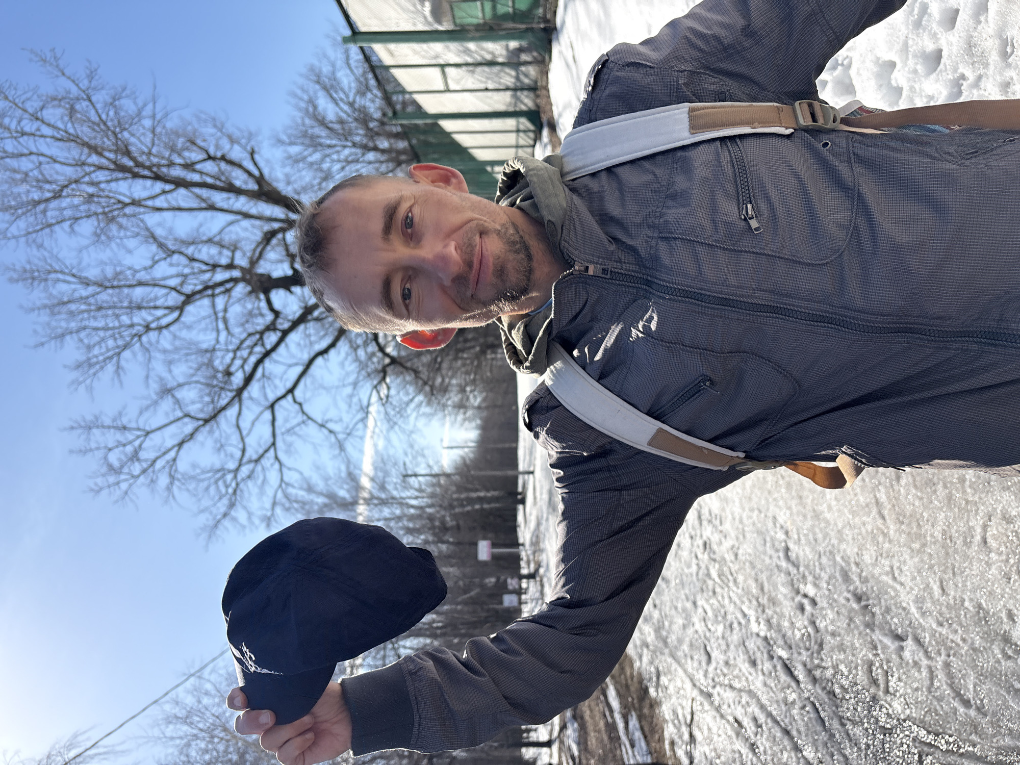 The image depicts a middle-aged man outdoors, holding a black baseball cap. He is wearing a grey-blue jacket with a visible zipper and hood, and white straps of what appears to be a backpack or harness are visible on his shoulders. The ground is covered with snow, and in the background, there are bare trees and a structure that appears to be a fence or enclosure. Based on the visual information, it's impossible to confirm the exact location within Ulyanovsk, Russia. More information would be needed to be able to identify the location.
