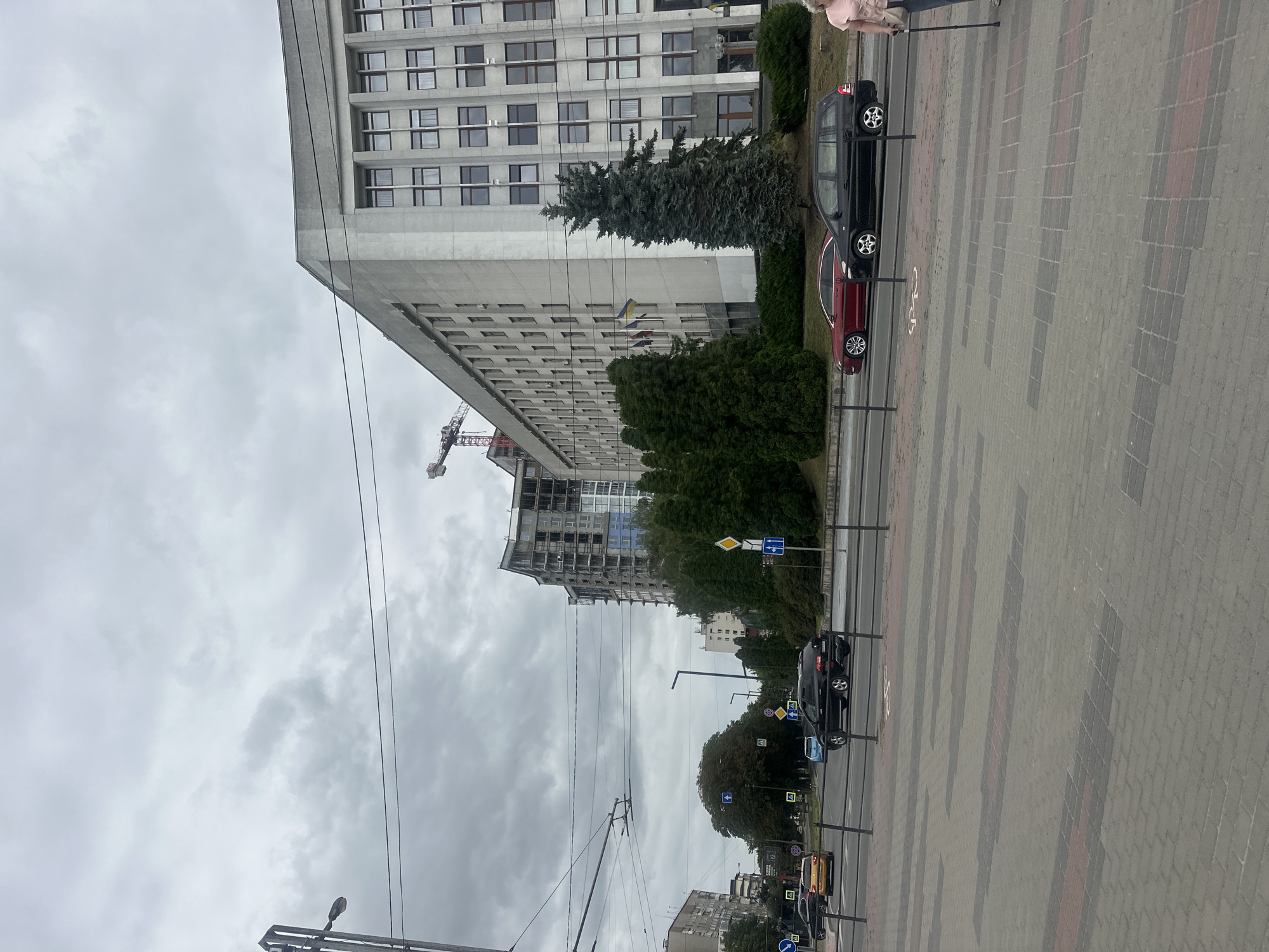 This is a wide-angle, eye-level, outdoor, daylight photograph taken on a cloudy day in Ternopil, Ukraine. The image depicts a street scene featuring a large, grey, rectangular, Soviet-era style building on the right side of the frame. This building has a repeating pattern of windows with dark frames and a slightly curved facade. In front of this building, there are several tall evergreen trees and manicured hedges. To the left of the grey building, there is a construction site with a tall building under development, complete with scaffolding and a crane. The street in the foreground shows a road with several cars, including a yellow sedan, a dark SUV, and a red sedan. The pavement in the foreground is made of grey and reddish-brown interlocking bricks, with a bicycle lane marking visible. A lamppost is present on the left side of the image, along with overhead power lines. In the far right, a woman is standing, holding a walking cane. Several traffic signs, including directional arrows and yield signs, are visible on the side of the road. The sky is overcast with dense grey clouds.