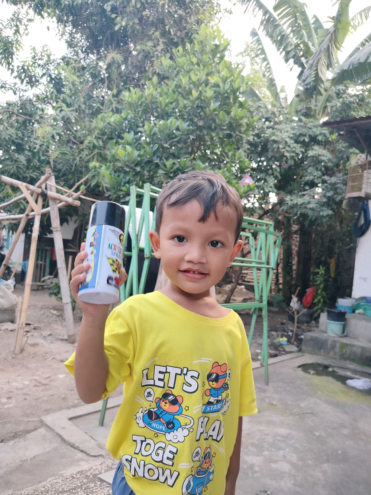 A young boy is standing outside in what appears to be a backyard area in Beloh, Indonesia. He is wearing a bright yellow t-shirt with cartoon graphics and blue jeans. The boy is holding a can of acrylic spray paint in his right hand, which is positioned at eye level with the can facing forward. He is looking directly at the camera with a slight smile. In the background, there are trees, bamboo structures, a green metal frame, and some scattered debris, suggesting a rural or semi-rural setting. The ground is dirt and concrete.