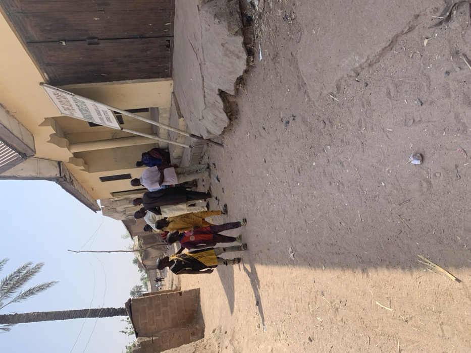 The image depicts an unpaved dirt street in Bojude, Nigeria, under clear, sunny daylight. On the left side of the street, three children walk towards the viewer; one wears a yellow and black sleeveless shirt, another a red and purple shirt, and the third a yellow top. To their right, along the dirt path, a crumpled white paper and several small sticks are visible.
On the right side of the street, a light-yellow building with a corrugated metal roof stands, featuring an entrance with steps and ornate columns. A large white sign with black text, supported by two metal poles, leans in front of this building, displaying "THE JODANIY... LAUNDRY & DRY CLEANING... ADAMAWA STATE." Adjacent to the sign, large wooden double doors are closed. A group of approximately seven adults stands near the building's entrance, with one individual seated on the steps.
In the background, on the far left, a tall palm tree is prominent against the clear sky, with other smaller buildings and distant figures visible along the dirt path. A thin wooden pole with attached wires extends upwards on the left side of the frame. The ground near the building exhibits a raised, cracked concrete platform.