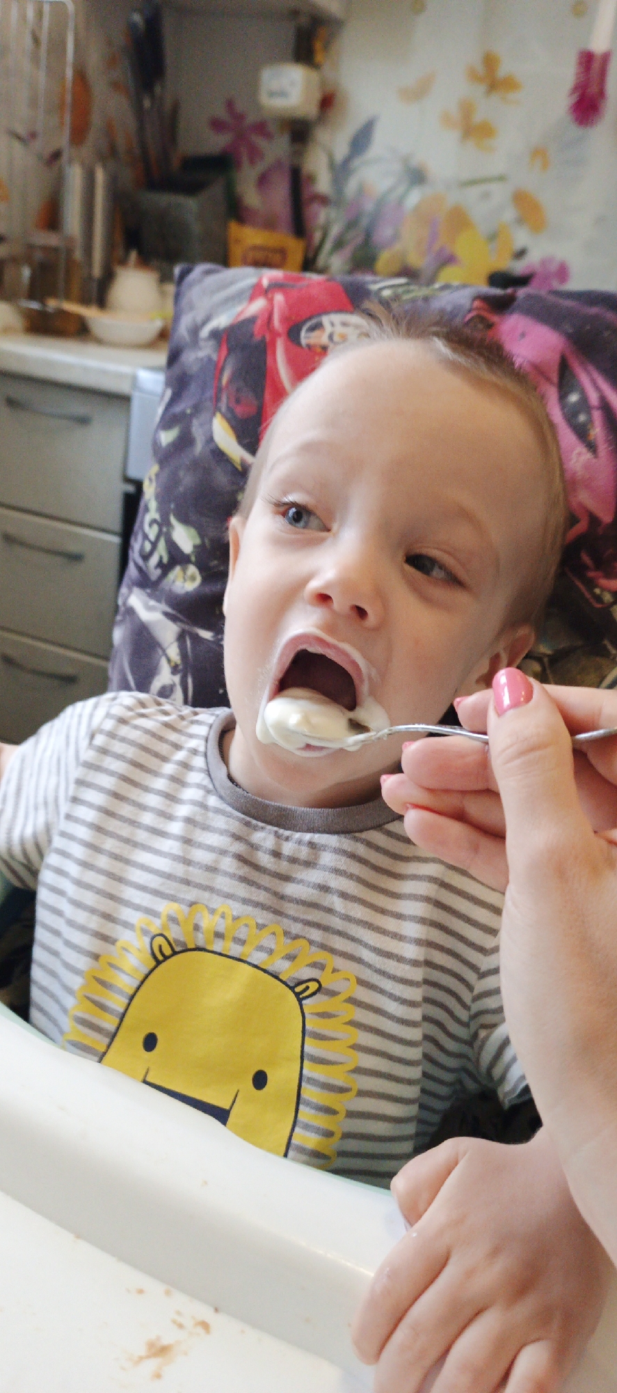 The image shows a close-up of a young child being fed by an adult indoors, set in a domestic environment, likely a kitchen or dining area in Chaikovsky, Russia.
The central subject is a fair-skinned child with light hair and blue eyes, positioned in a white high chair. The child's mouth is wide open, actively receiving a spoonful of a white, creamy food, possibly yogurt or puree, from an adult. Some of the food is visibly smeared around the child's mouth. The adult's hand, adorned with pink nail polish, holds a metal spoon directly in front of the child.
The child is wearing a grey and white striped short-sleeved t-shirt with a large yellow lion graphic printed on the chest. Behind the child's head, a pillow with a distinct red racing car design on a dark background is visible. The background of the scene is blurred but reveals typical household elements: grey kitchen cabinets with drawer pulls on the left, and a patterned wall or curtain featuring floral and abstract designs in shades of pink, yellow, and green. Various kitchen items, including what appears to be a metal appliance and small bowls, are partially visible on a counter behind the child. On the white tray of the high chair, there are a few reddish-brown smudges, suggesting prior spills. The lighting indicates it is daytime. No discernible text is visible in the image. The child's expression suggests concentration or anticipation during the feeding process.