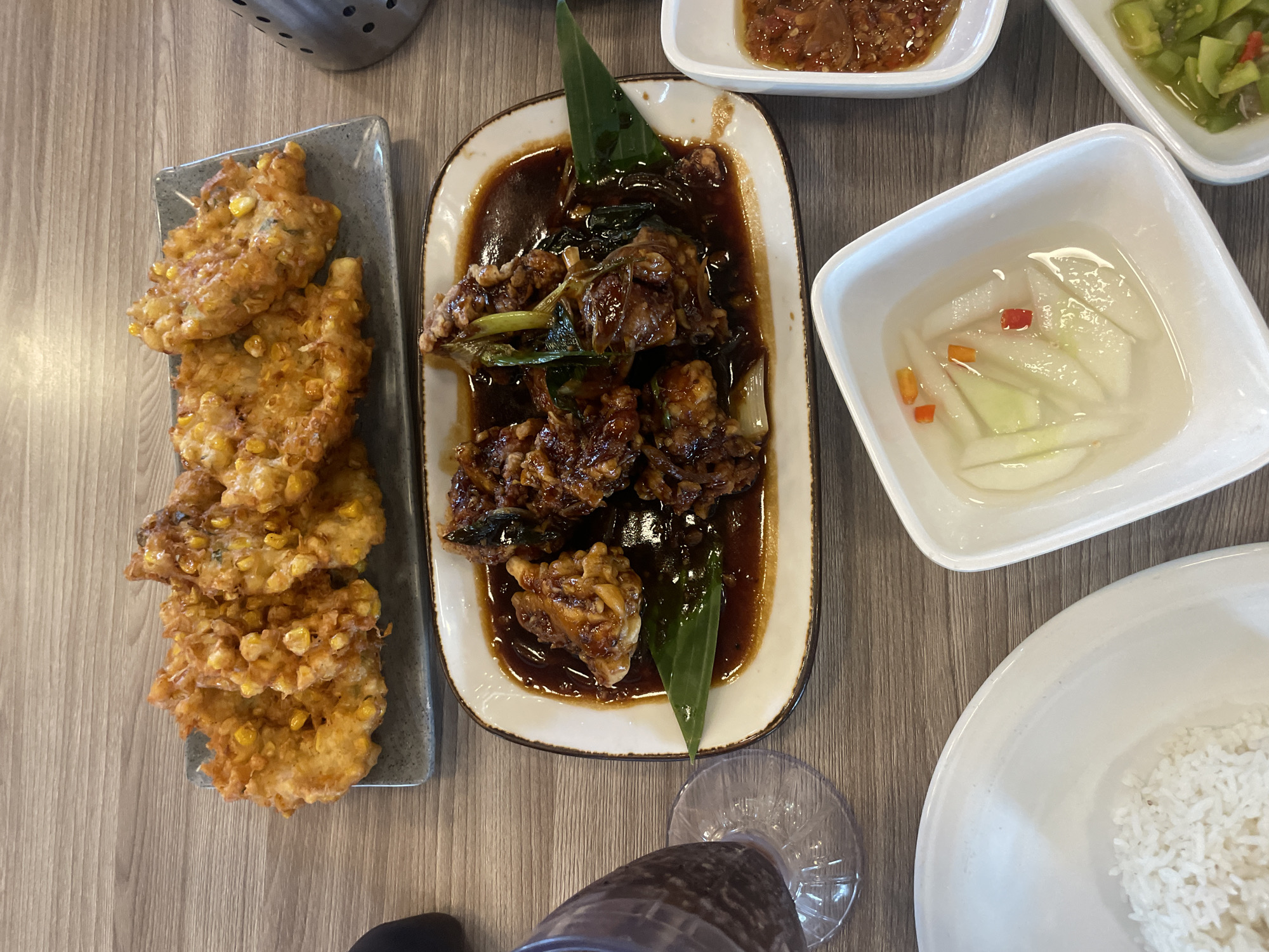 A overhead shot captures a variety of dishes on a wooden table, likely at a restaurant in Manado, Indonesia. The arrangement includes a rectangular grey platter filled with golden-brown corn fritters, each visibly containing whole kernels of corn. To the right of the fritters, a portion of a metallic shaker with perforated sides is visible.
Dominating the center of the image is a white oval platter holding pieces of dark, glossy food coated in a rich sauce, garnished with green leaves and stalks. To the left of this platter, a clear glass with a dark liquid sits on a coaster, partially obscuring the wooden surface.
In the foreground, a white square bowl contains a medley of thinly sliced white vegetables, possibly cucumbers or radishes, interspersed with small pieces of red and orange chili. Another white square bowl to the right holds a similar pickle, this time with green vegetables, likely tomatoes, and red chili. Further to the right, a small white dish contains a reddish condiment, possibly a sambal. Finally, a large white bowl on the left is filled with fluffy white rice. The overall scene suggests a meal in progress, with multiple dishes prepared for sharing.