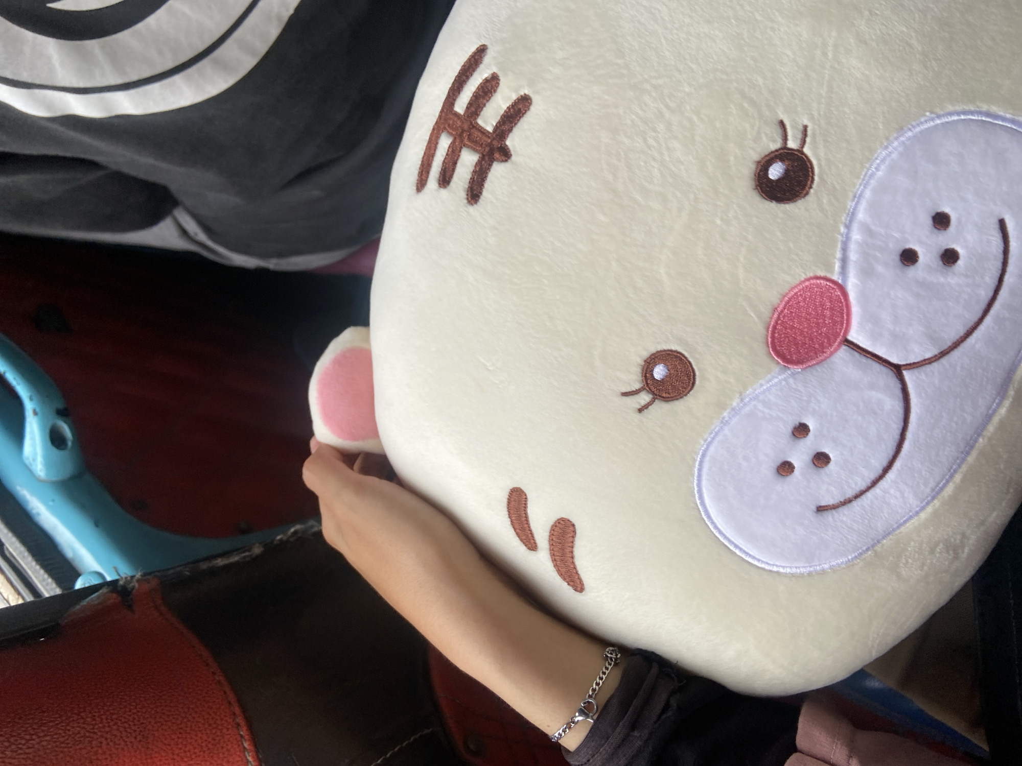 A person's arm, adorned with a silver chain bracelet, rests on a plush, cream-colored stuffed animal with embroidered brown tiger stripes and facial features. The stuffed animal has a pink nose and light purple accents around its face. The background shows the interior of a vehicle with red and blue seats. The scene appears to be within a public transportation vehicle in Pangkalan Kerinci, Indonesia.