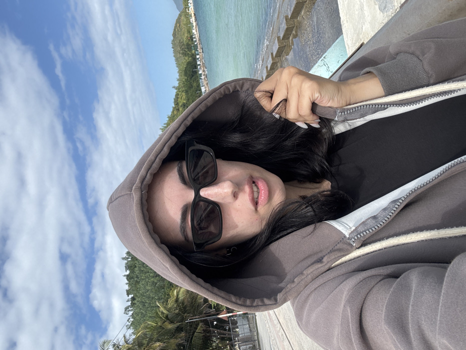 A portrait-style image, rotated 90 degrees counter-clockwise, features a person, likely a young woman, looking directly at the camera. She wears a grey or taupe hooded zip-up sweatshirt over a black inner shirt. Her dark, wavy hair spills out from under the hood. She is also wearing dark, rectangular-framed sunglasses that cover her eyes, with her well-defined eyebrows visible above. Her lips are slightly parted. One hand, with long, light-colored nails, is visible on the right side of the frame, touching her hair near the hood.
The background is divided between a clear blue sky with scattered white clouds on the upper left and a scenic coastal landscape on the lower right. The tranquil, turquoise-blue sea meets concrete or rock structures along the shore. Further in the distance, lush green hills and scattered buildings, possibly resorts or houses, are visible among trees, including palm trees. The scene is brightly lit by natural daylight, suggesting good weather. The image conveys a relaxed mood against the beautiful backdrop of Ninh Hòa, Vietnam. There is no visible text in the image.