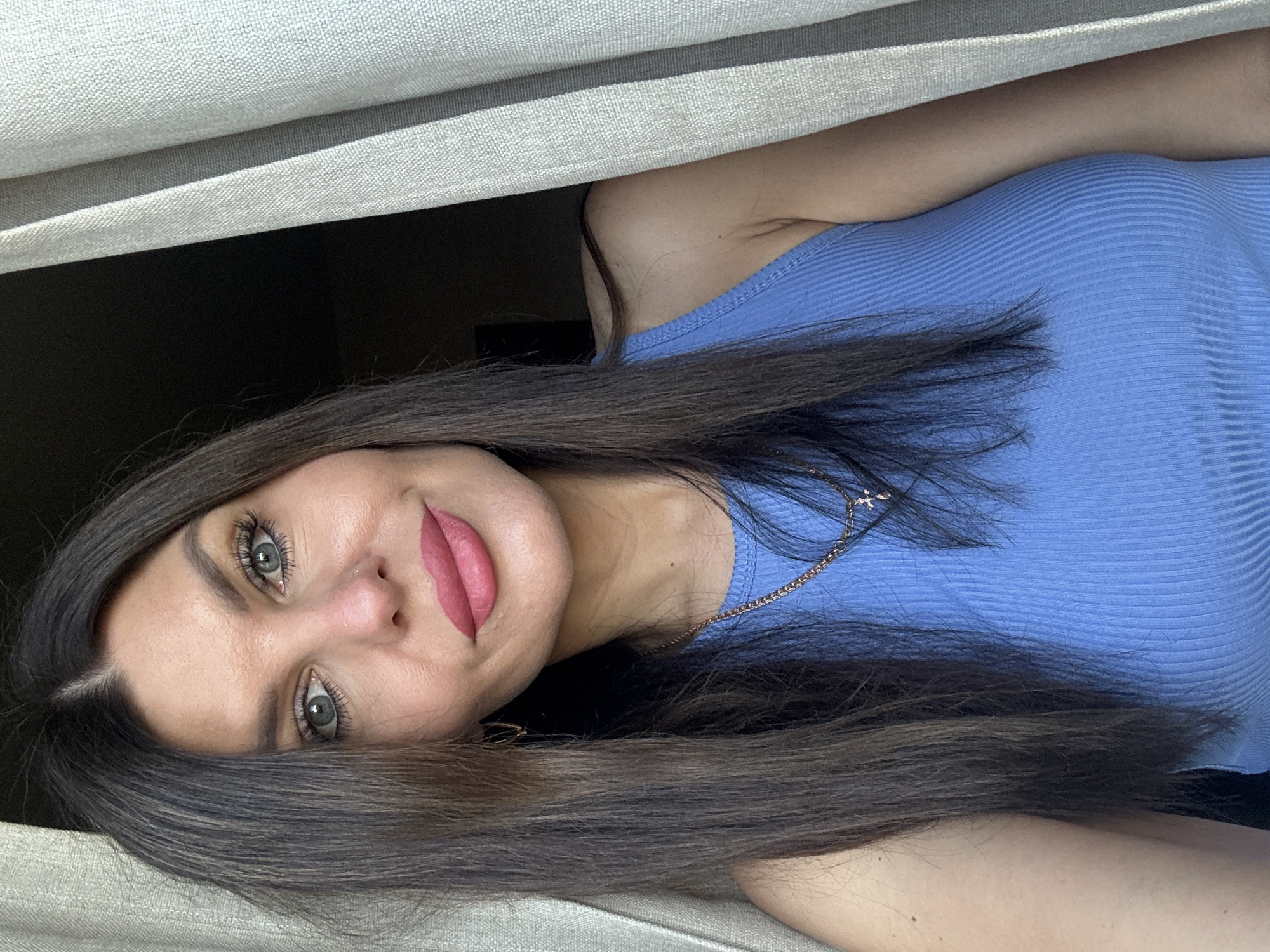 A woman with long dark hair and blue eyes, wearing a blue tank top and a gold necklace, looks directly at the camera. She is positioned in front of a light-colored curtain, with a dark background on the left. The image is a close-up shot of her face and upper body. There is no identifiable information to suggest the location is Chaikovsky, Russia.