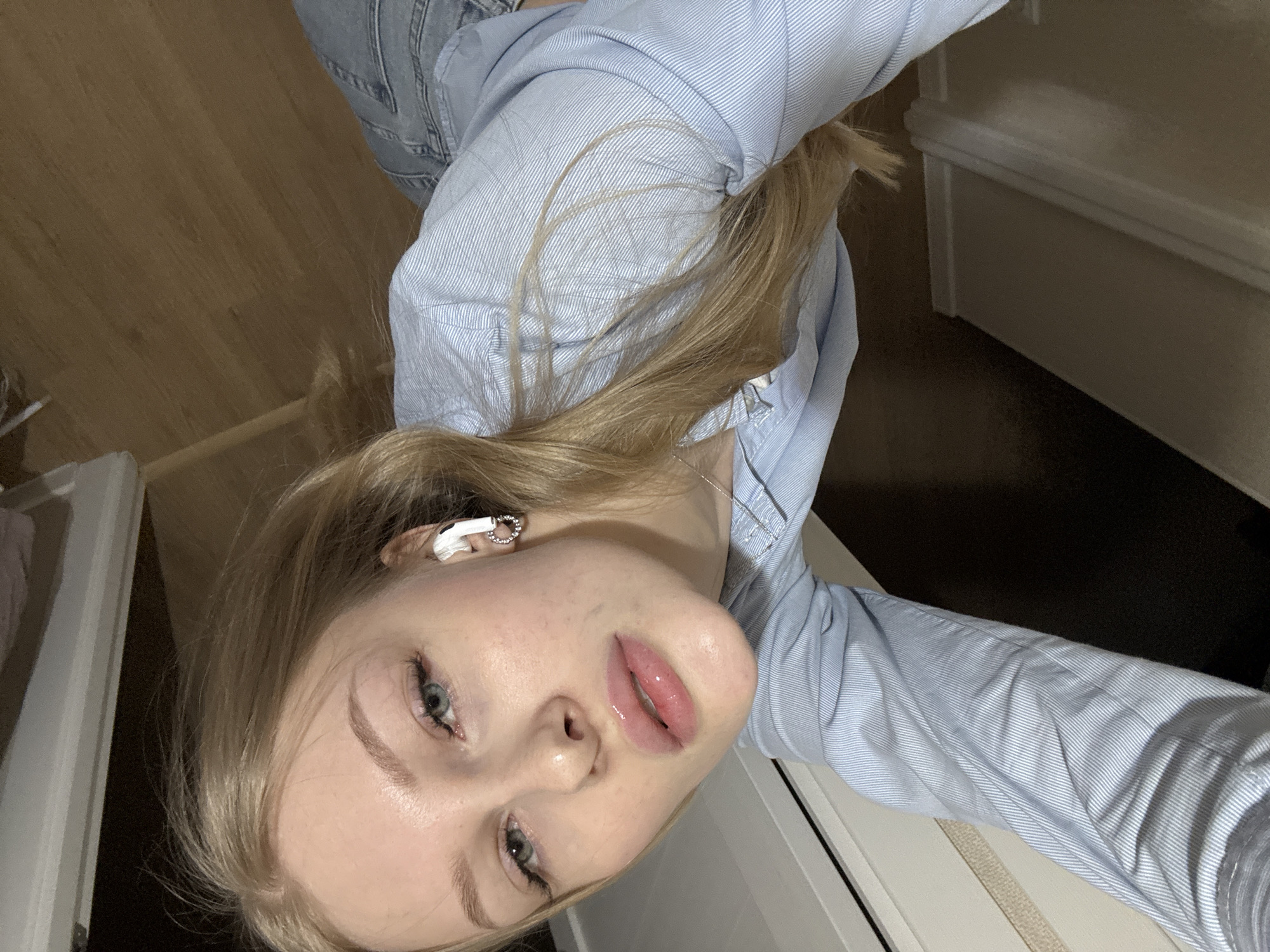 A young woman is captured in a tilted, close-up selfie taken indoors, likely in Perm, Russia. She has long, straight blonde hair, fair skin, and striking light blue eyes, gazing directly into the camera. Her full lips are slightly parted, showcasing a glossy finish. She has well-defined eyebrows and appears to be wearing mascara. In her right ear, a white earbud with a distinctive silver, circular detail is visible.
She is dressed in a light blue shirt with fine white stripes, which is partially covered by what appears to be a denim garment, visible in the upper left of the frame. Her blonde hair spills over her shoulder and arm. The background suggests an indoor setting, with a light brown wooden floor visible to the left and a white wall or door frame element towards the right and bottom, indicative of a residential or private space. The lighting is soft and even, highlighting her features. The overall mood is casual and natural. No visible text is present in the image.
