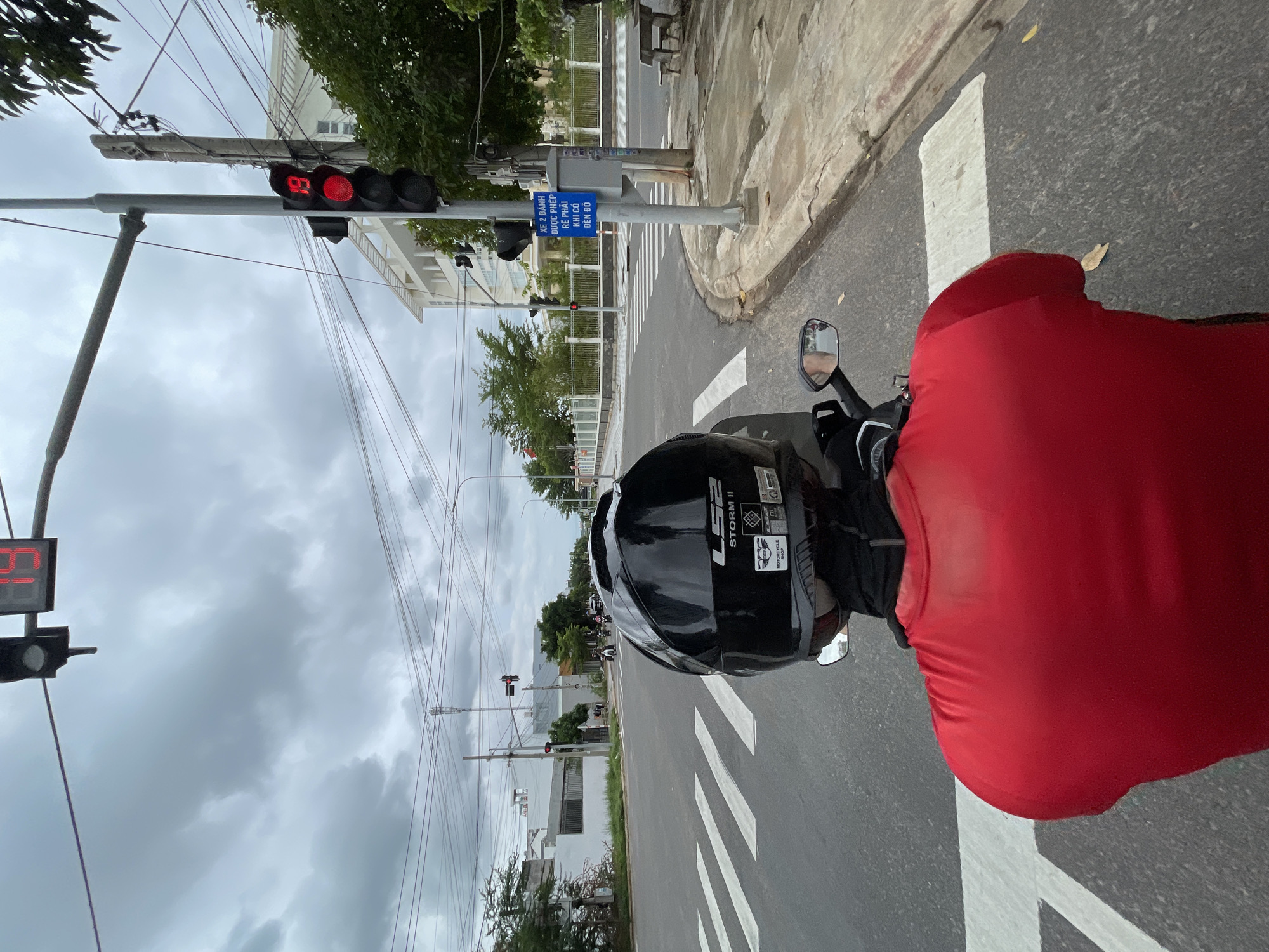 This is a first-person perspective image taken from a motorcycle at an intersection in Phu Thuy Ward, Vietnam. The rider, wearing a red shirt and a black helmet, is stopped at a crosswalk. A traffic light is visible, showing a red signal with a countdown timer displaying "19". Other motorcycles are present at the intersection, suggesting it is a busy junction. The environment includes roads, sidewalks, buildings, trees, and overhead power lines, under a cloudy sky. A sign in Vietnamese is mounted on a pole next to the traffic light.