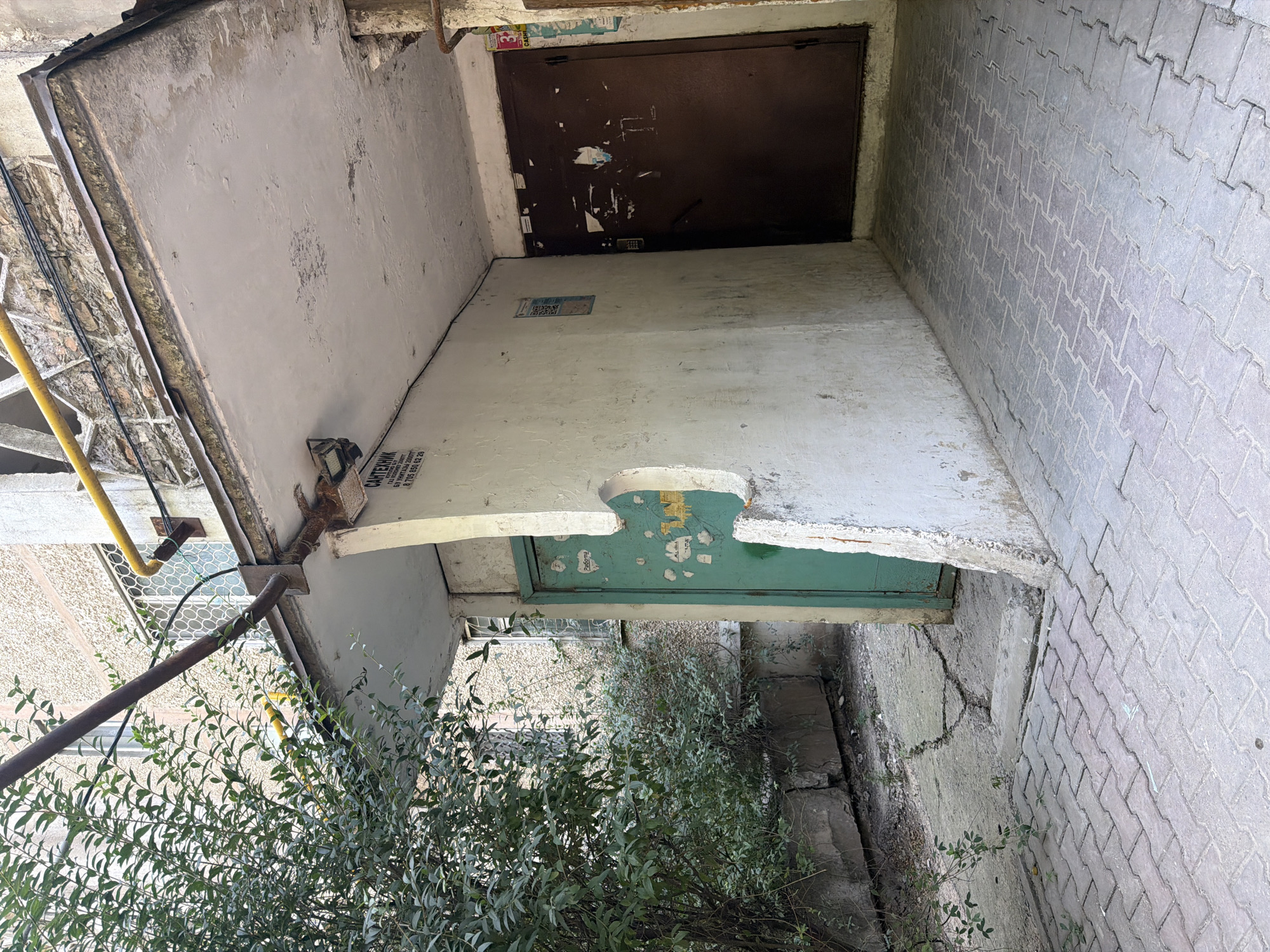 The image depicts an exterior view of an apartment building entrance in Shymkent, Kazakhstan. In the foreground, interlocking paving stones form a walkway. To the left, a dense green bush with small leaves grows next to a concrete wall. Ahead, a teal-colored door is visible, adorned with several stickers, one of which appears to be a heart shape. The door is set into a weathered, textured wall that has a cut-out decorative element. To the right of this door, a dark brown metal door is present, also covered in various stickers and peeling paper. Above the entrance, a concrete overhang shows signs of wear and tear, with exposed rebar and peeling paint. A rusty metal pipe runs horizontally across the top of the frame, supported by brackets attached to the building. Another yellow pipe runs diagonally above it. The building's facade, visible in the background, is textured and light-colored, with barred windows. The overall scene suggests an older, somewhat neglected urban environment.